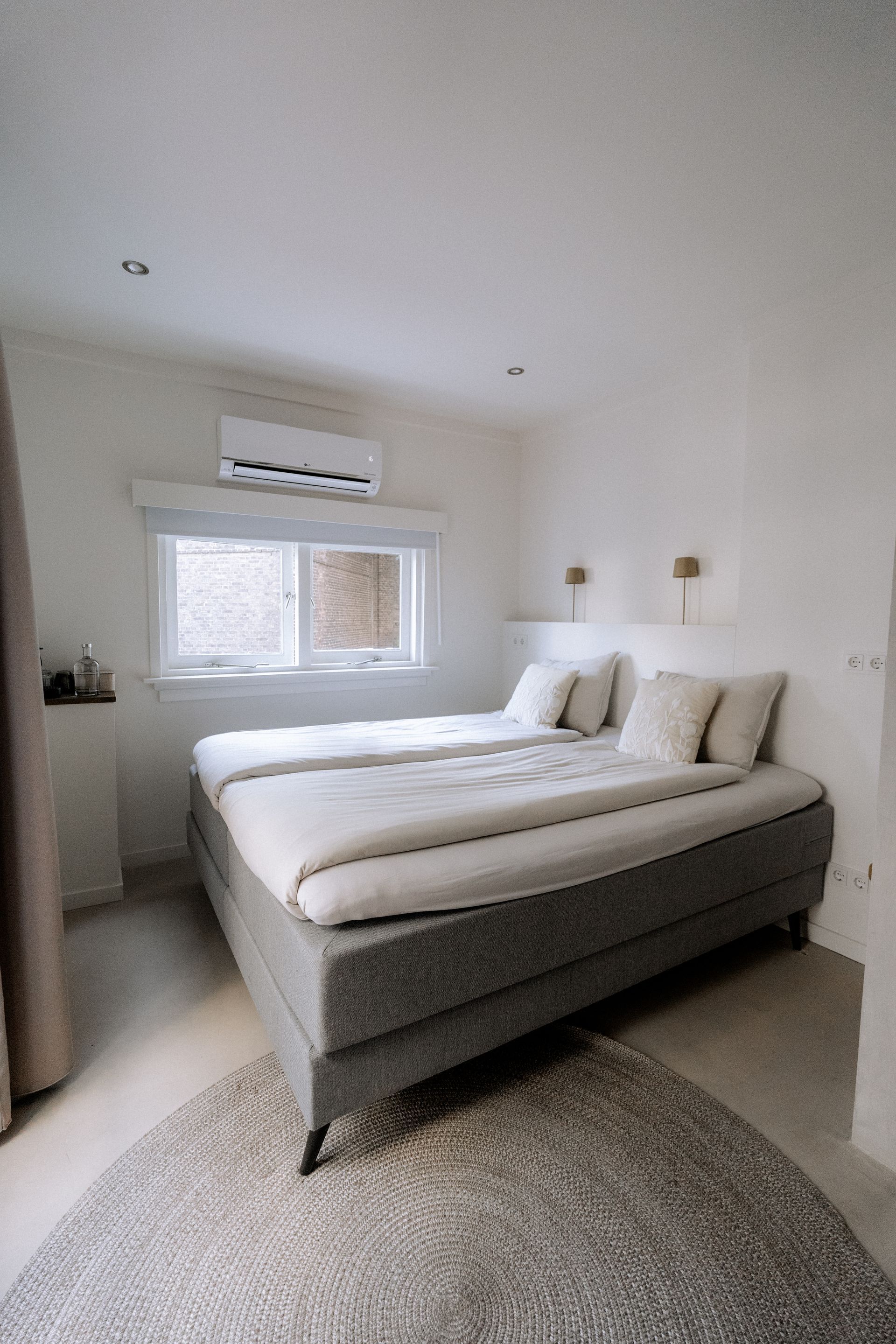 Slaapkamer met een tweepersoonsbed, grijs matras, raam en klein vloerkleed. Witte muren en airconditioning.