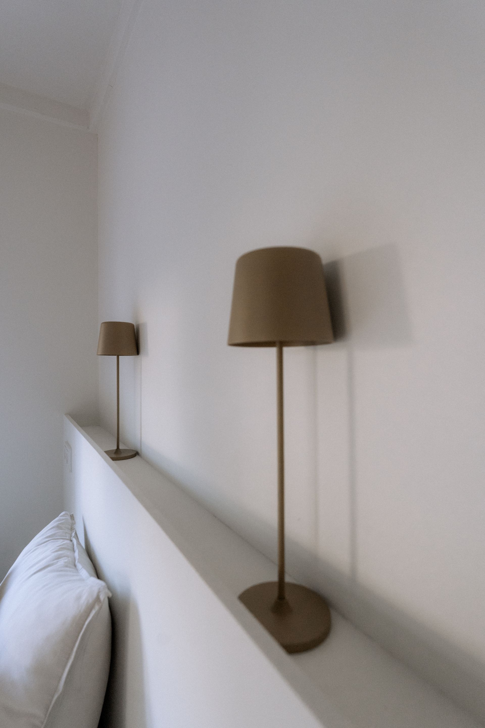 Twee lampen met bruine kappen staan op een witte plank boven een bed. Op het bed is een kussen zichtbaar.