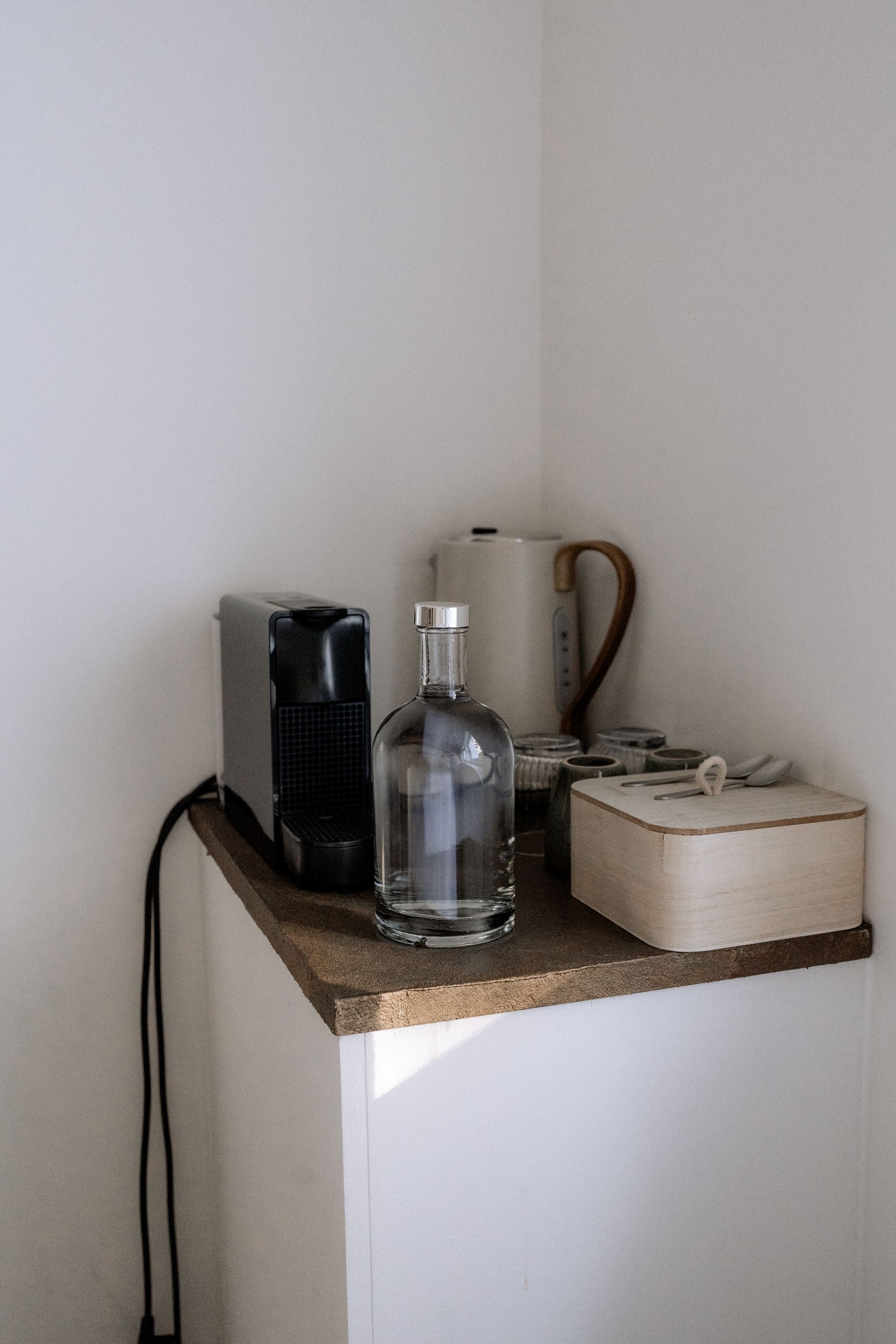Koffiestation met een koffiezetapparaat, fles, waterkoker en houten doos op een plank in een witte hoek.