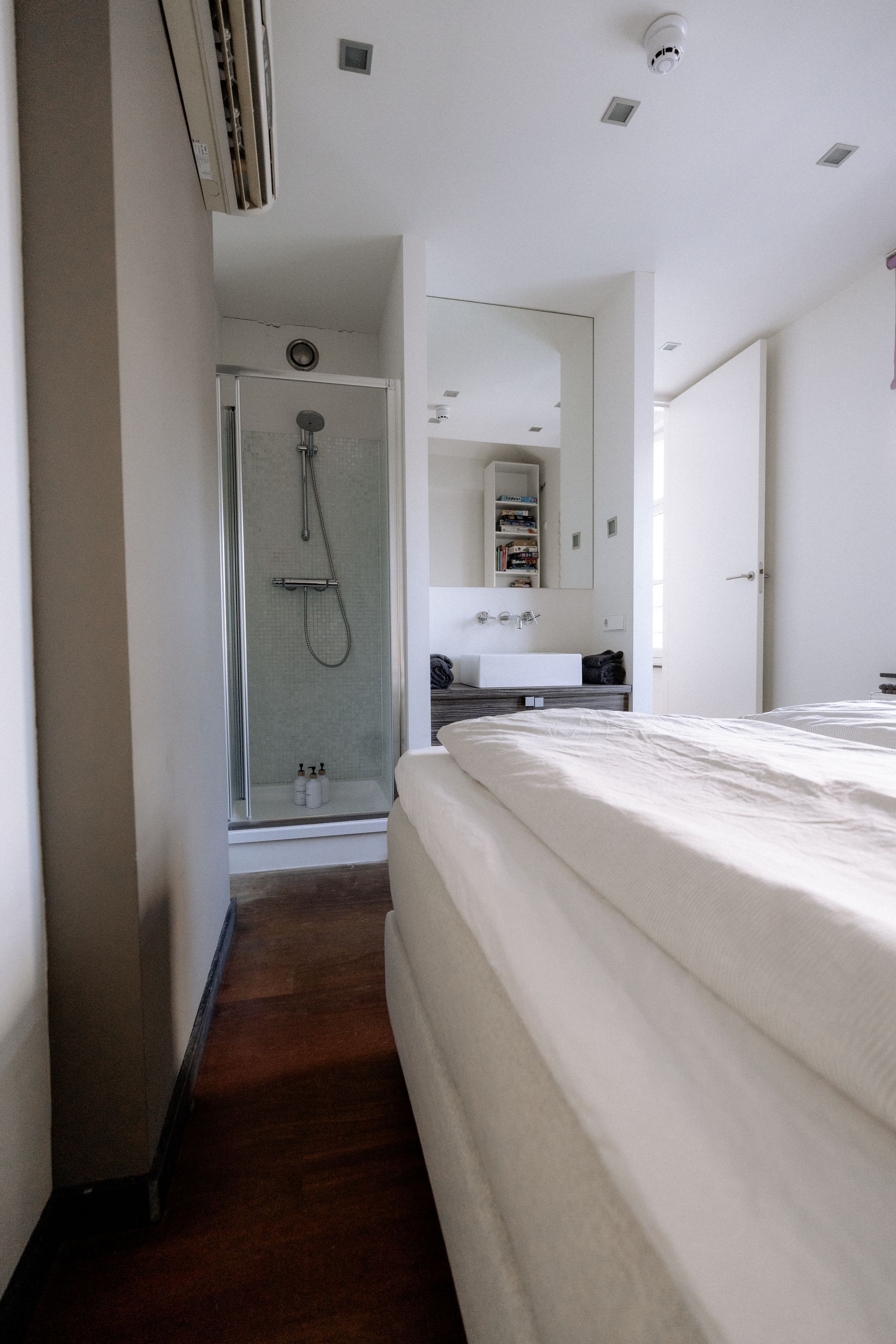 Slaapkamer met bed, douche en wastafel. Witte en bruine tinten, smalle ruimte, heldere verlichting.