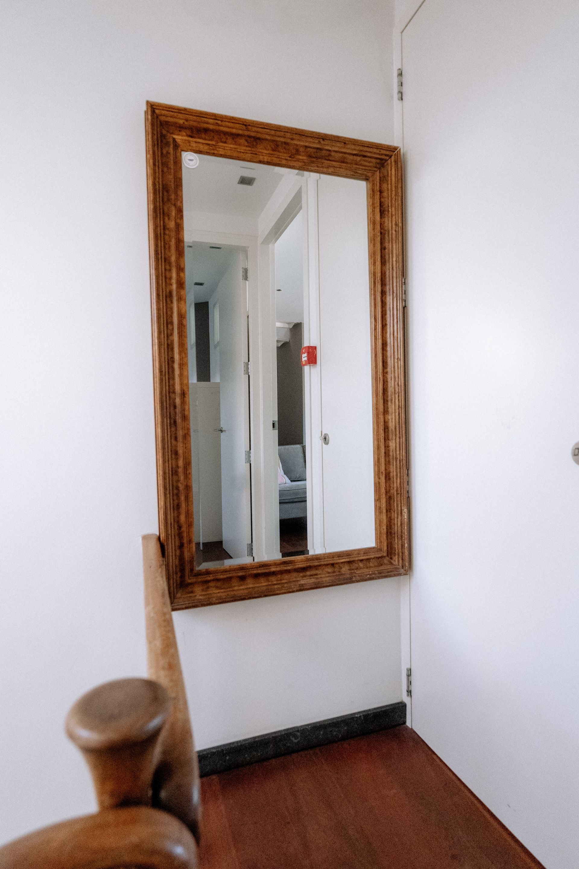Een spiegel met houten frame aan een witte muur. Op de voorgrond staat een houten stoel.