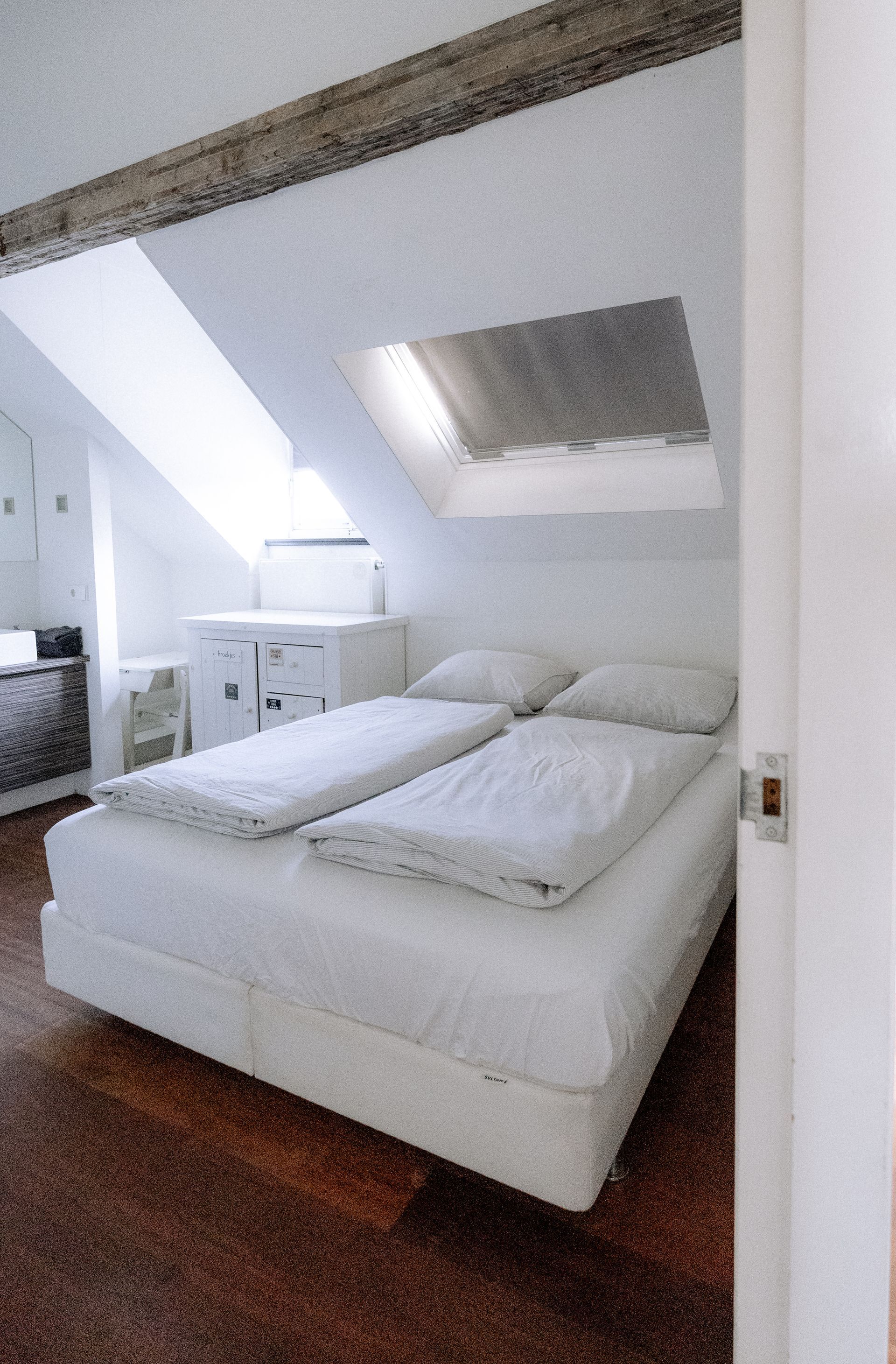 Slaapkamer met wit bed, schuin plafond met dakraam, houten vloer.