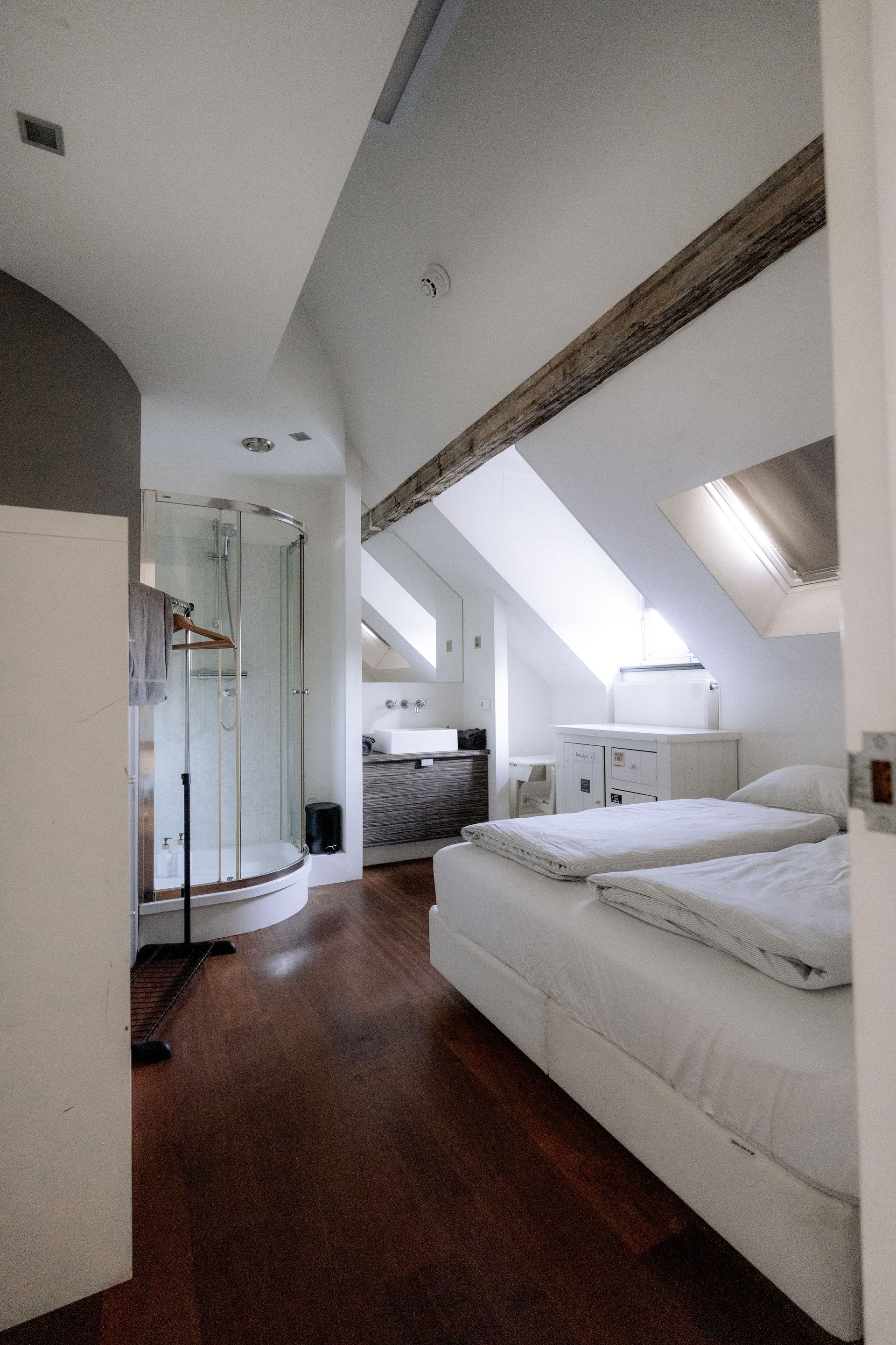 Slaapkamer met schuin plafond, houten balken, glazen douche, twee witte bedden en donkere houten vloeren.