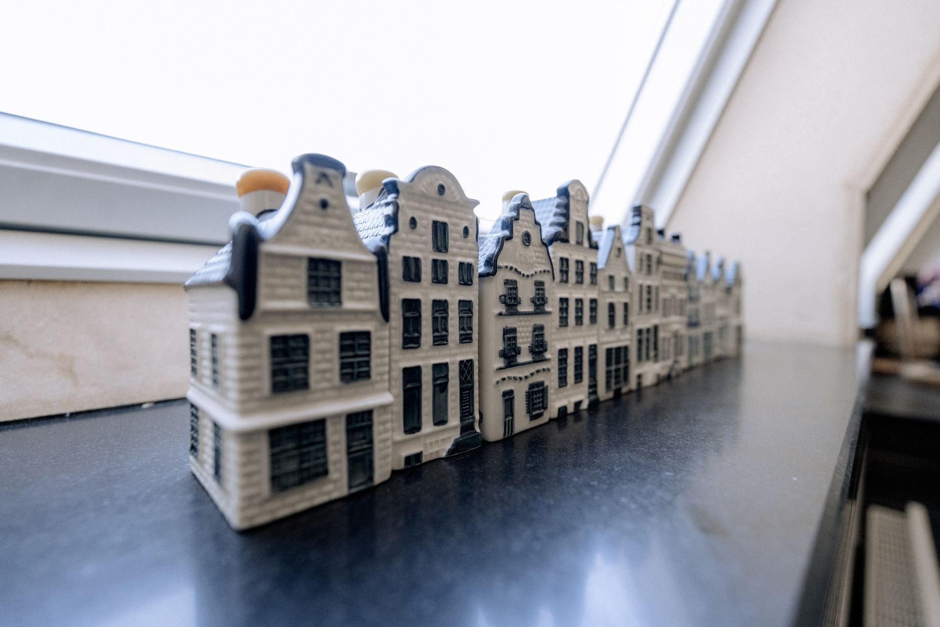 Een rij miniatuur keramische Amsterdamse grachtenpanden op een reflecterende donkere ondergrond bij een raam.