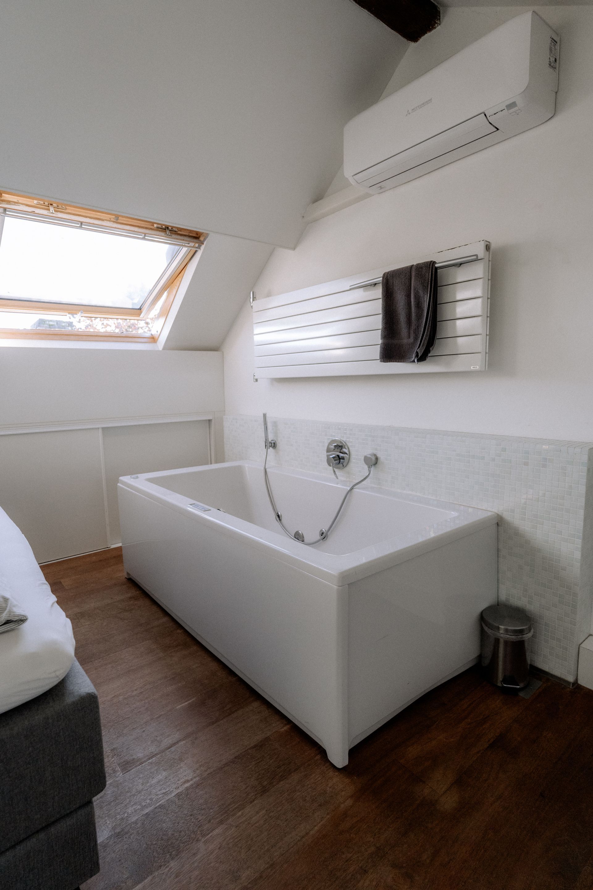 Badkamer met ligbad, dakraam en handdoekverwarmer. Witte muren en houten vloer.