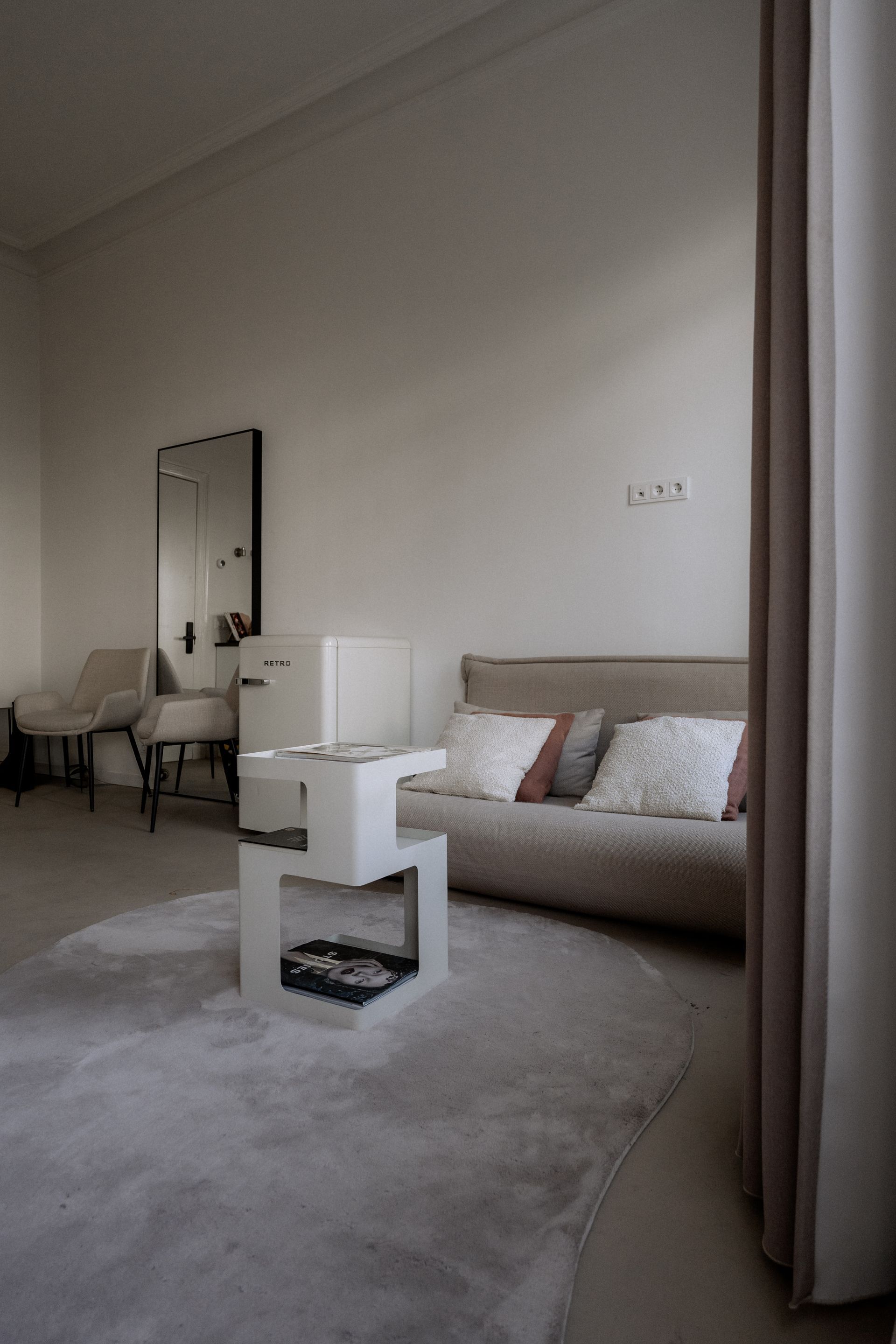Een kamer in neutrale tinten met een bank, een witte tafel, een vloerkleed, een spiegel en gordijnen.