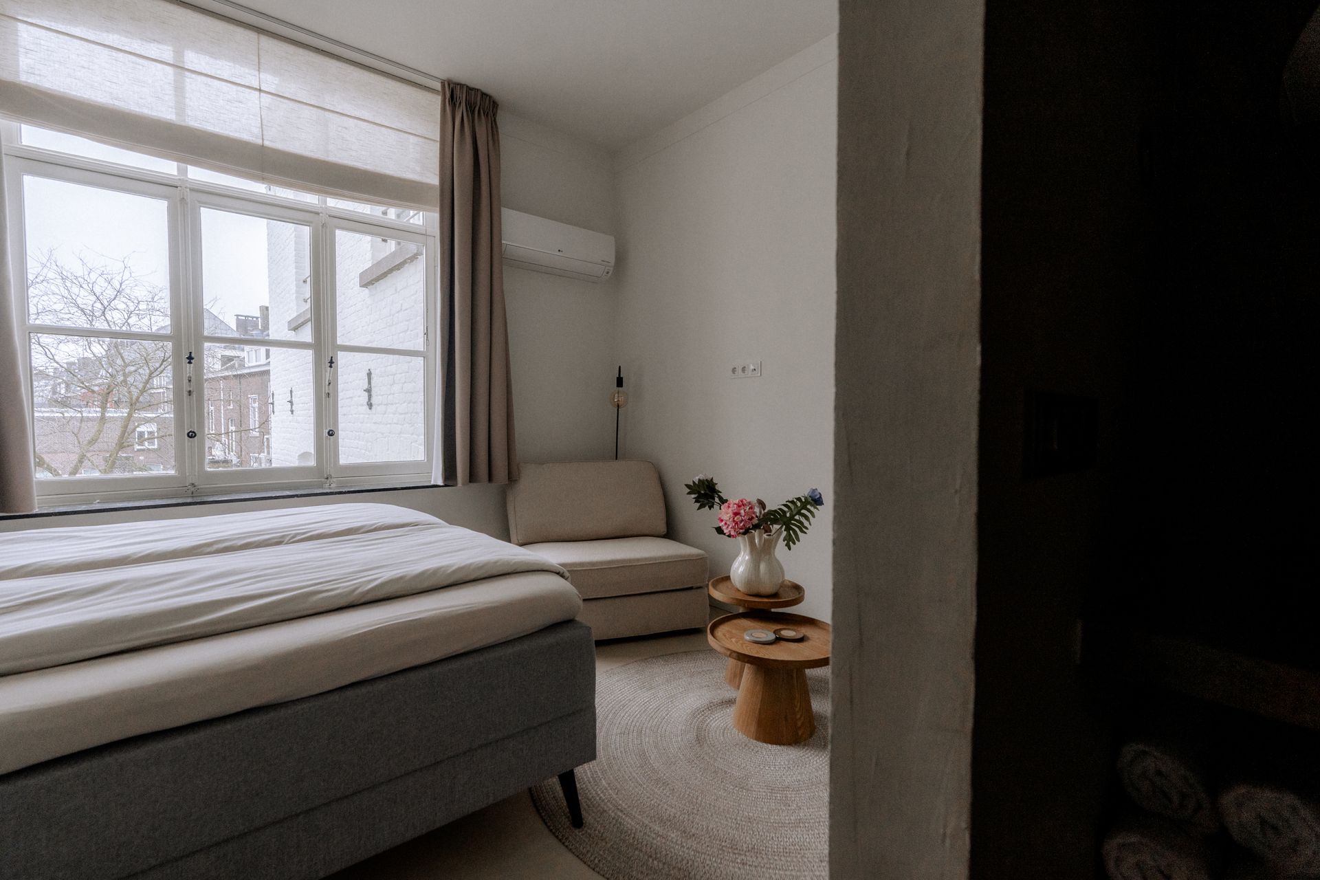 Slaapkamerinterieur met bed, stoel, tafel, raam en bloemstuk.