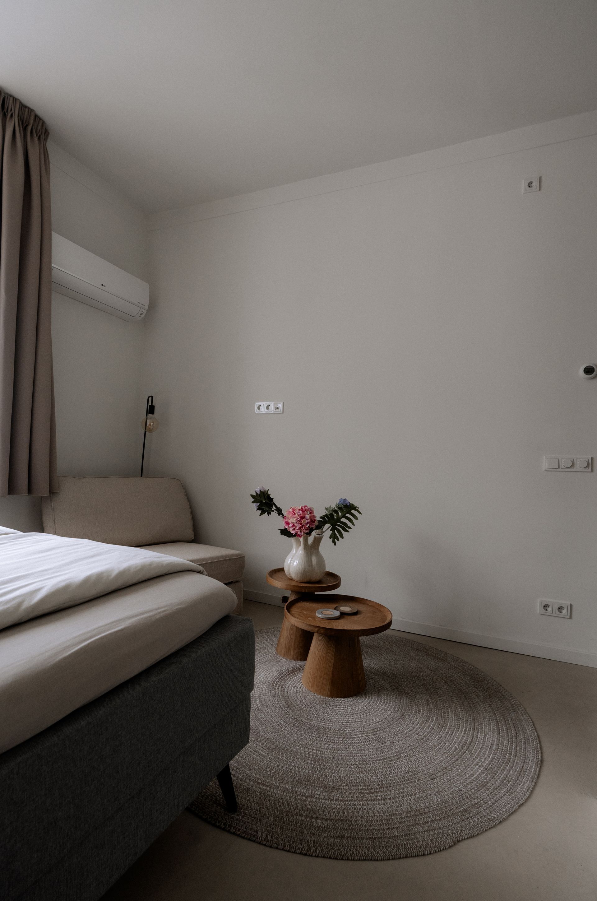 Slaapkamerinterieur: bed, neutrale muren, rond vloerkleed, kleine houten tafeltjes met bloemen.