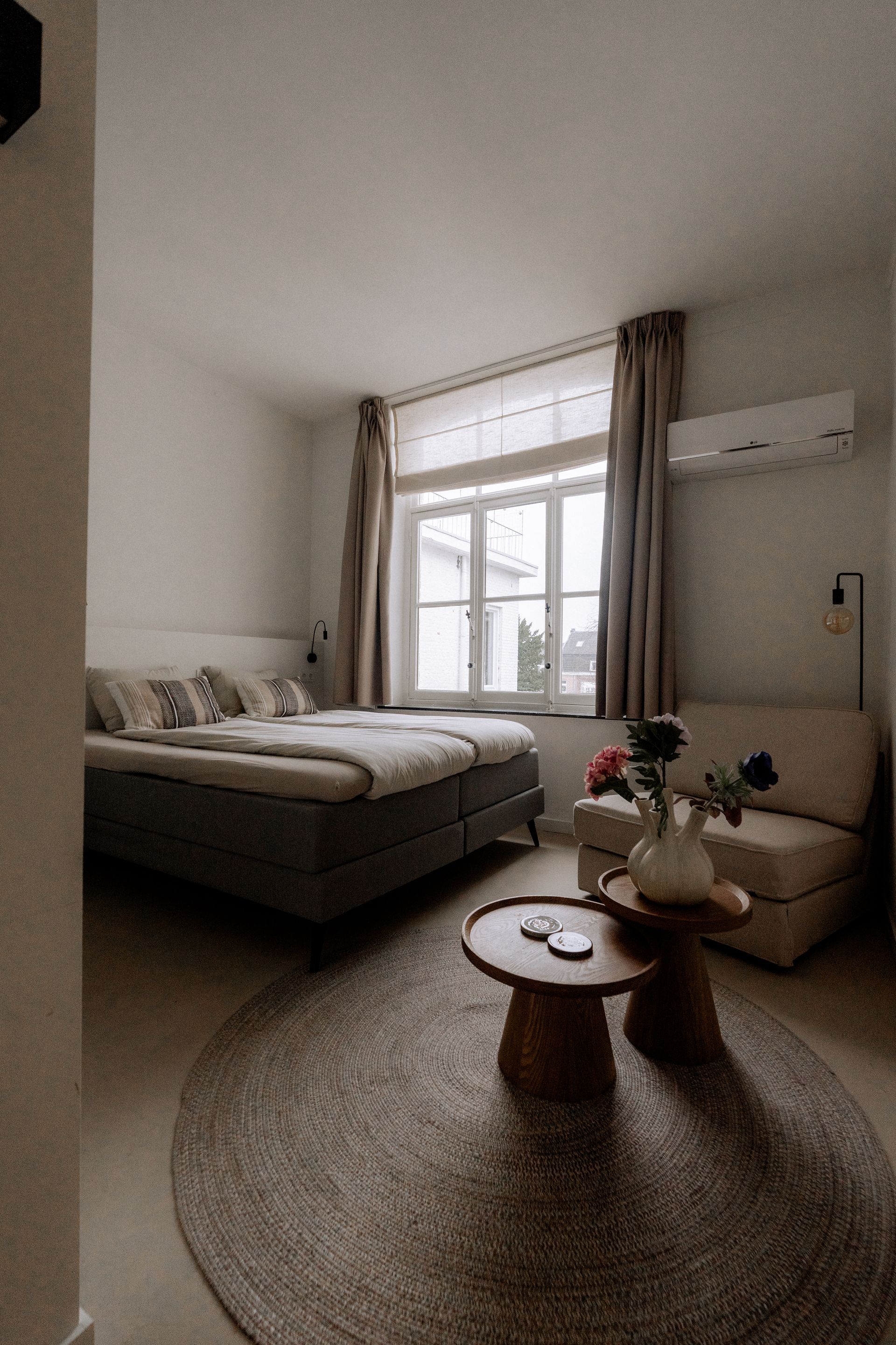 Slaapkamer met een tweepersoonsbed, zithoek, rond vloerkleed en raam met gordijnen.
