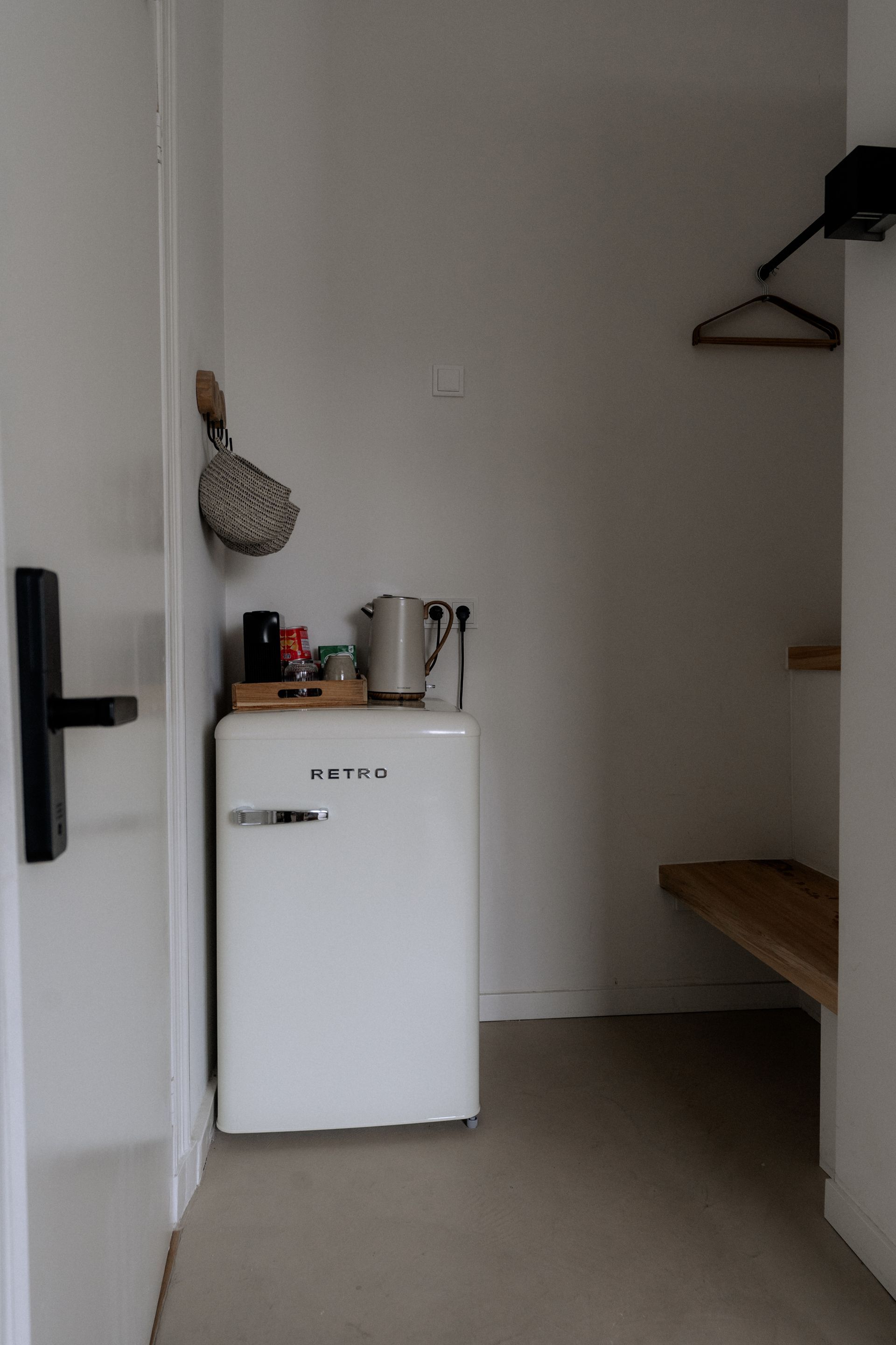 Een witte minikoelkast en een waterkoker in een kleine hal. Aan de rechterwand hangen een houten plank en een kledinghanger.