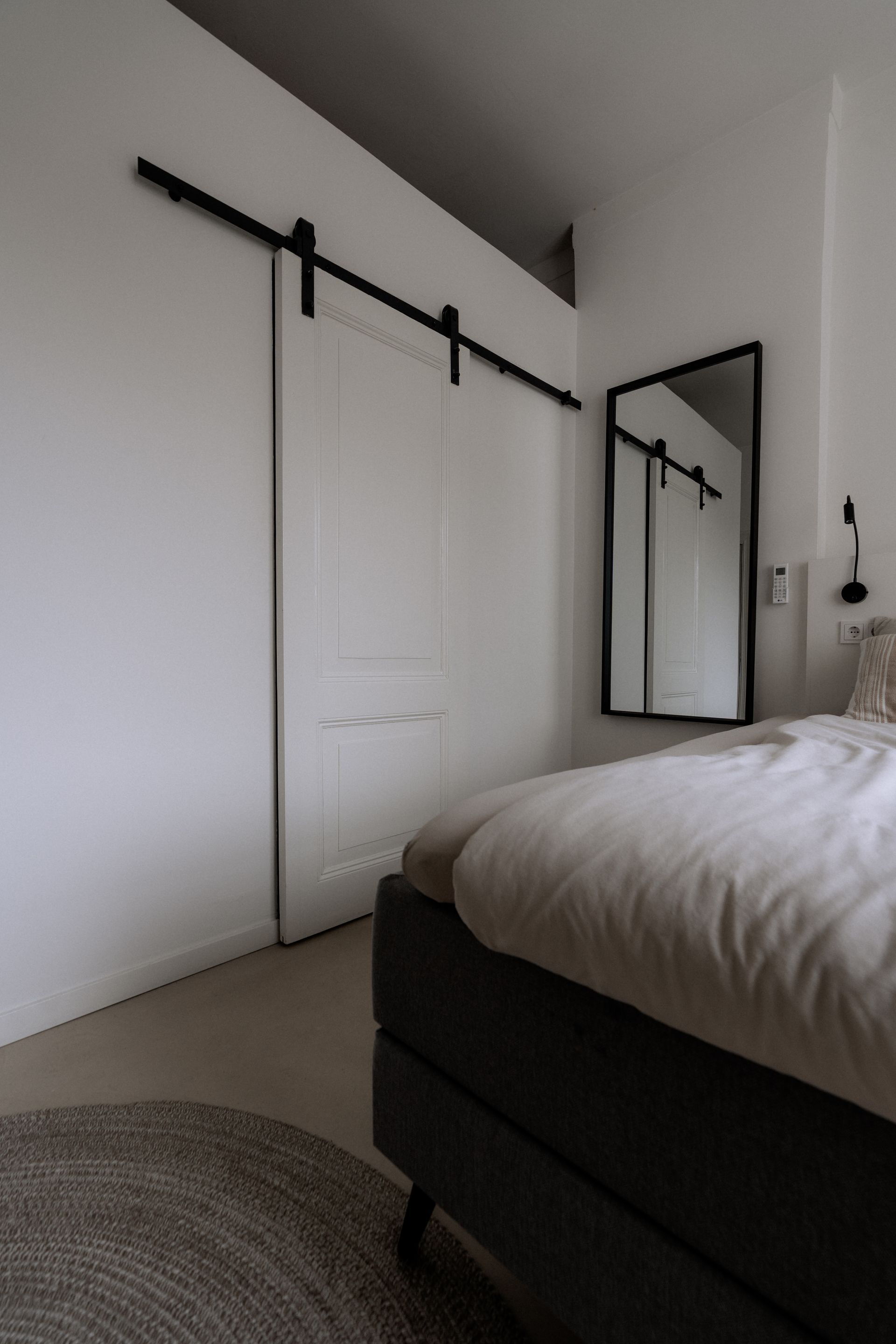 Witte slaapkamer met schuifdeur in boerderijstijl en spiegel. Bed op de voorgrond, vloerkleed linksonder.