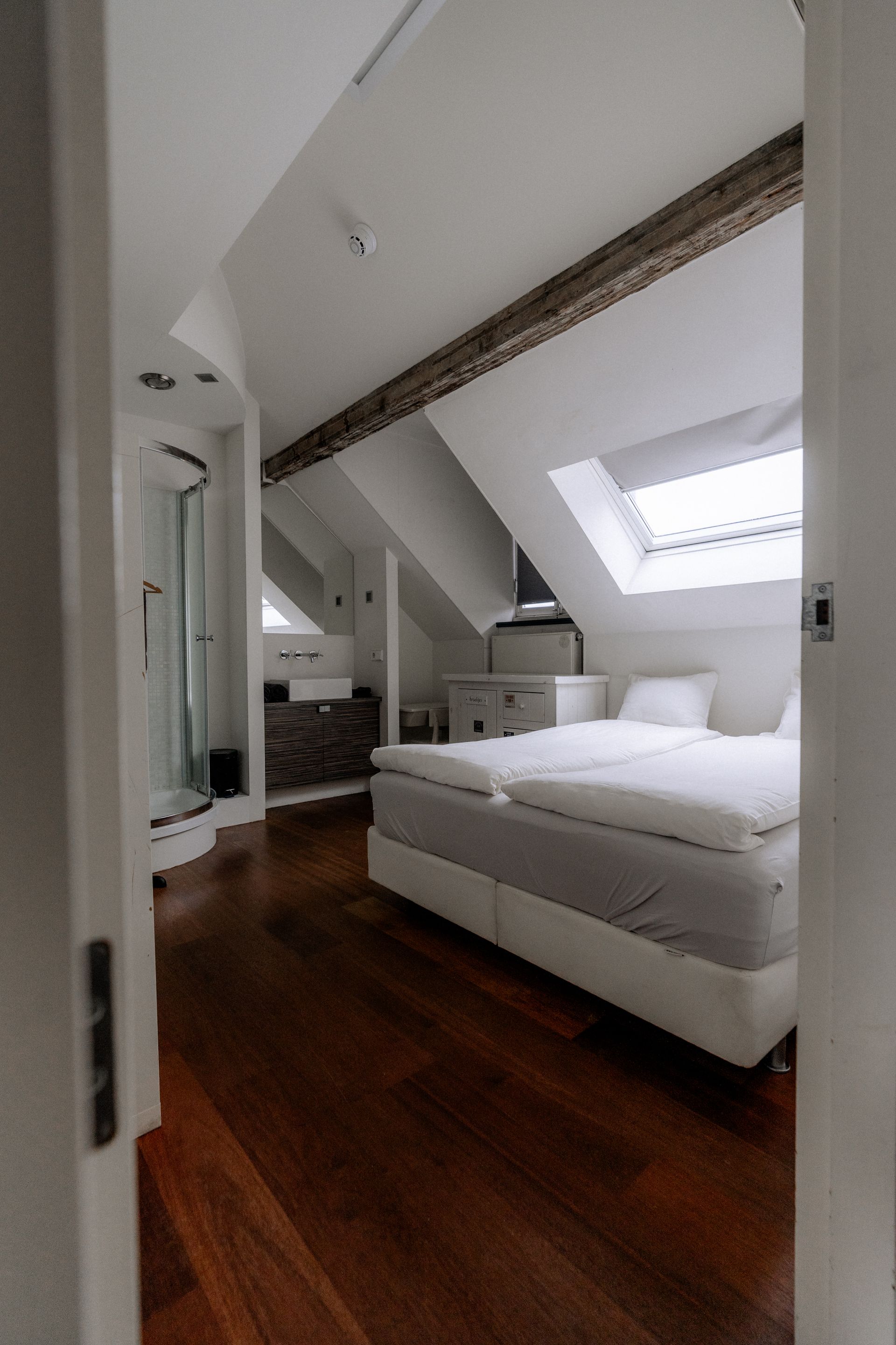 Slaapkamer met houten vloer, wit bed, dakraam en open badkamer.