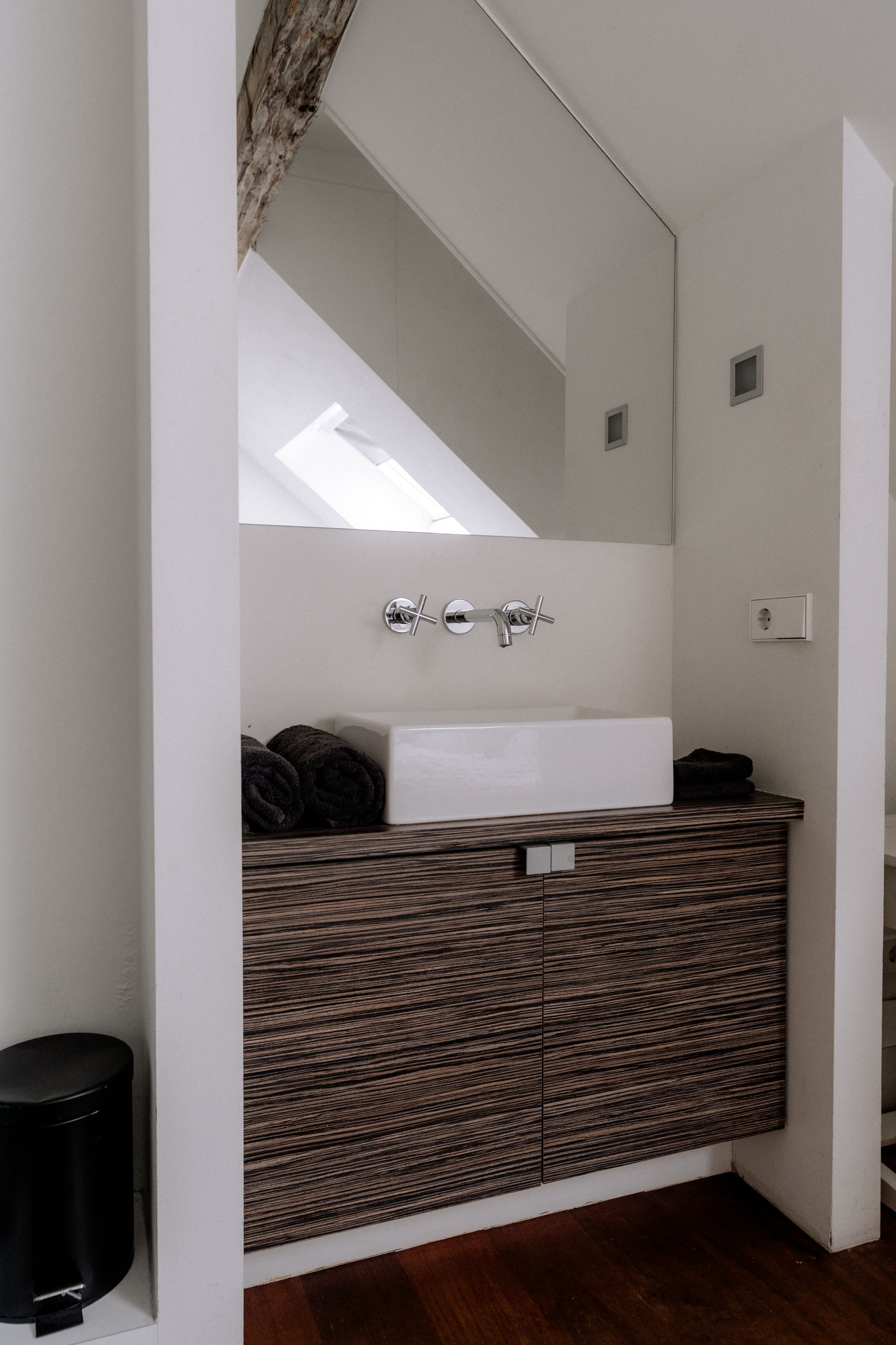 Badkamer met geweven kast, witte wastafel, grote spiegel en zwarte prullenbak.