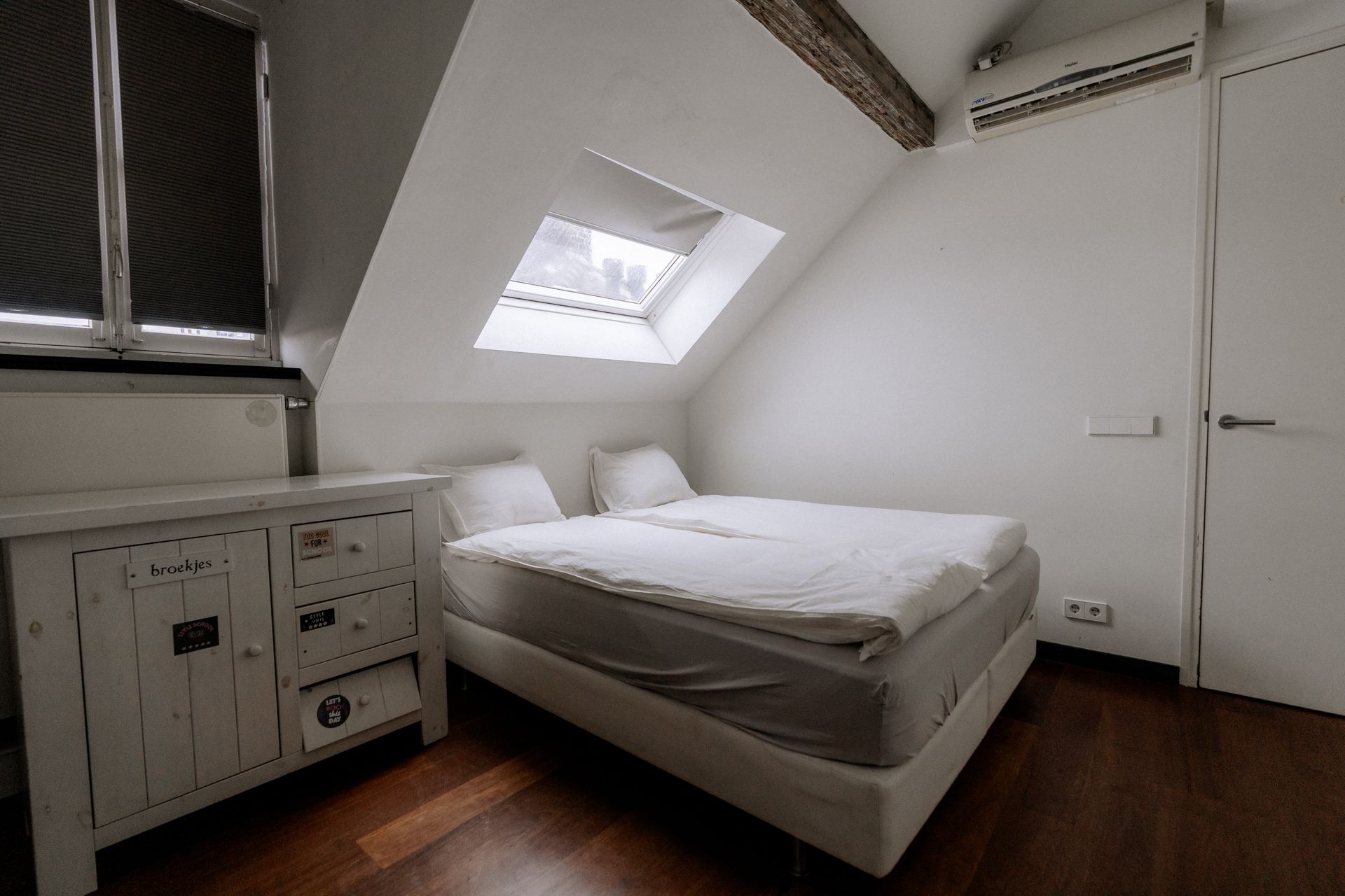 Slaapkamer met witte muren, dakraam, bed, commode en donkere houten vloer.