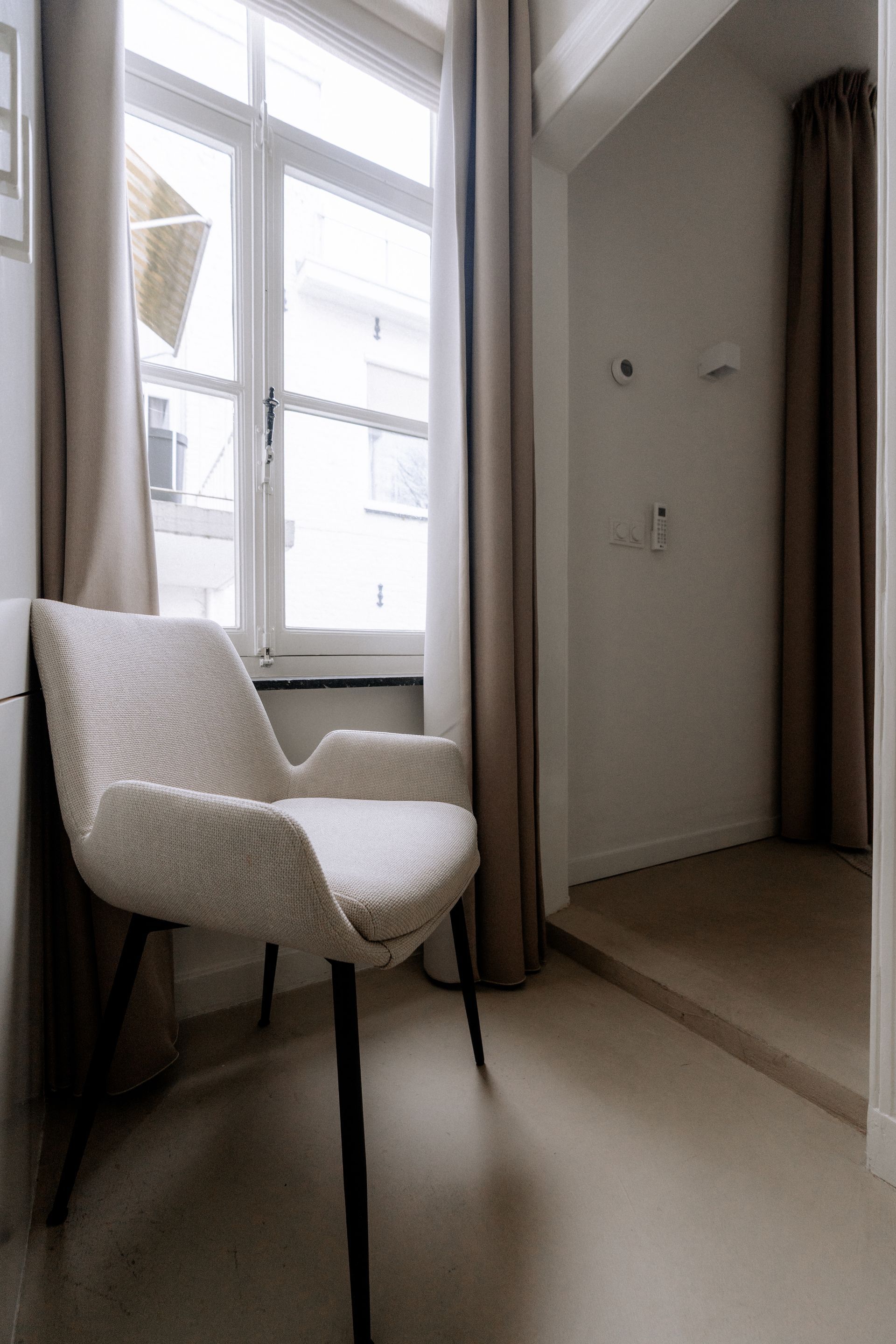 Crèmekleurige stoel bij het raam met beige gordijnen, in een kamer met lichtgekleurde vloeren.