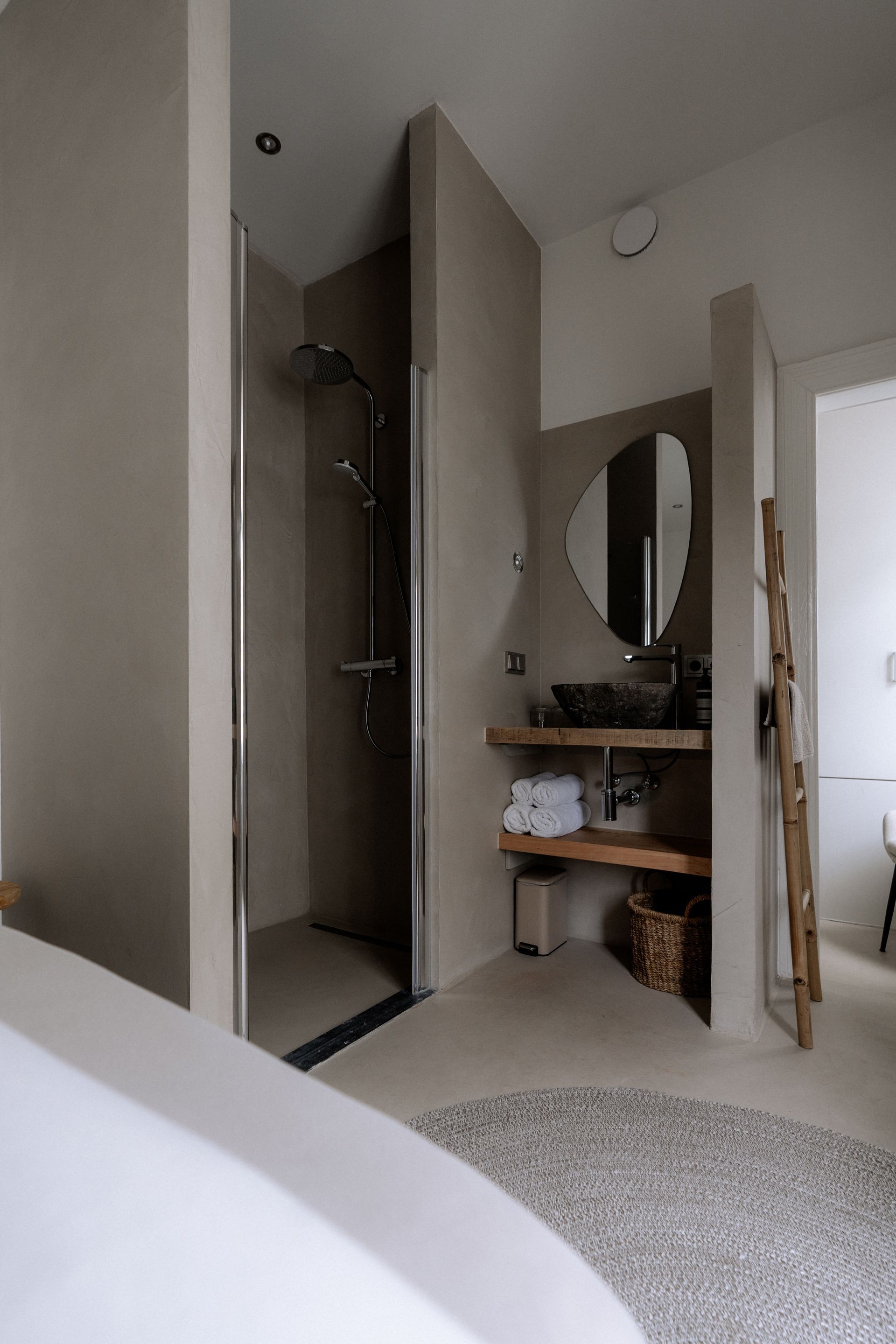 Badkamer met inloopdouche, wastafelmeubel met ronde spiegel en een klein vloerkleedje. Neutrale tinten.
