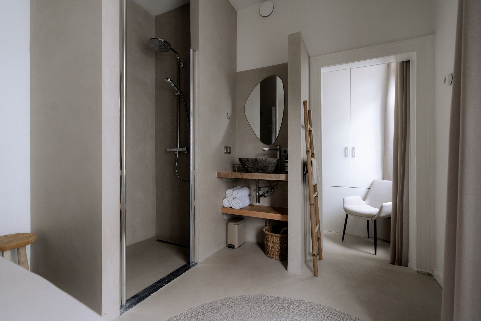 Badkamer met inloopdouche, wastafel en een glimp van een slaapkamer met een stoel. Neutrale tinten en modern design.