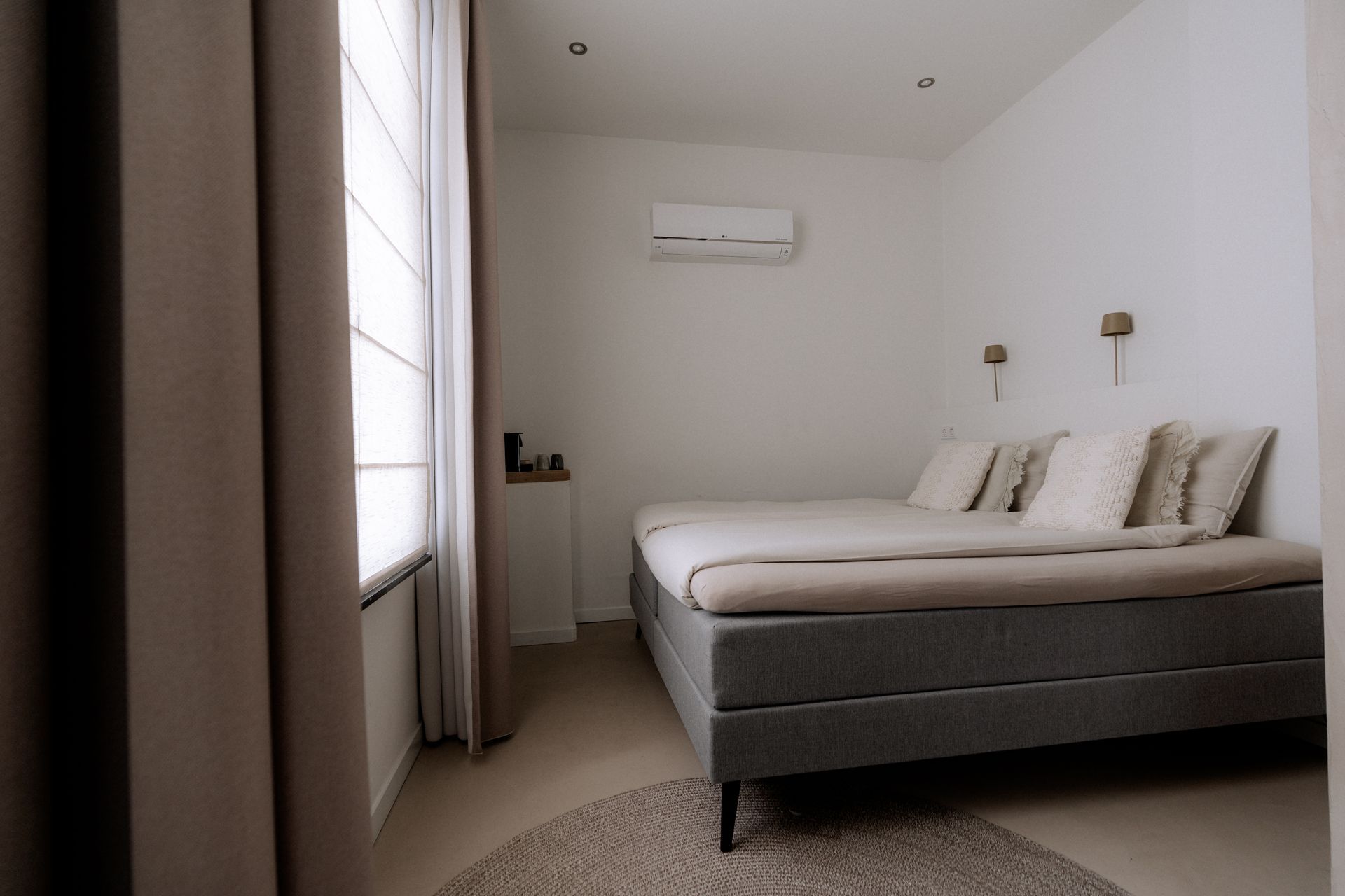Slaapkamer met een grijs bed, witte muren, een klein vloerkleed en een raam met beige gordijnen.