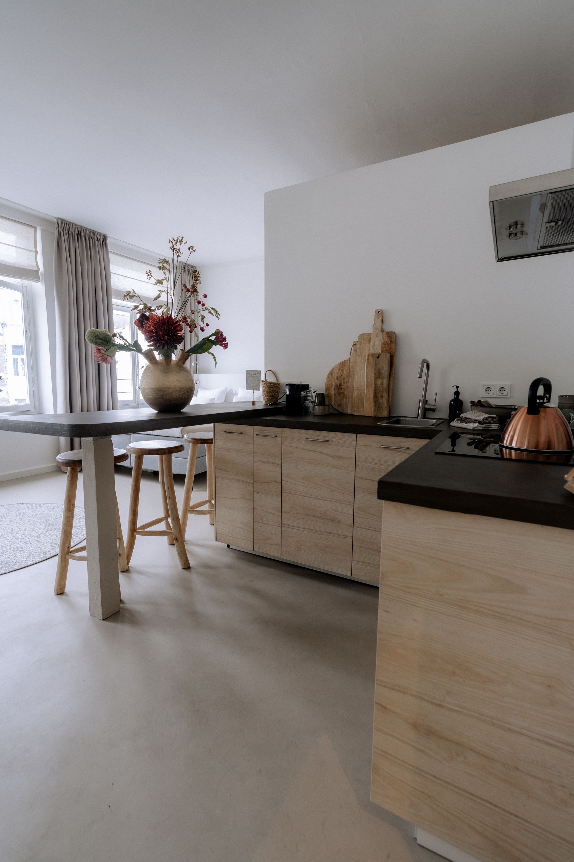 Moderne keuken met lichte houten kasten, donkere werkbladen en een eettafel met bloemen.