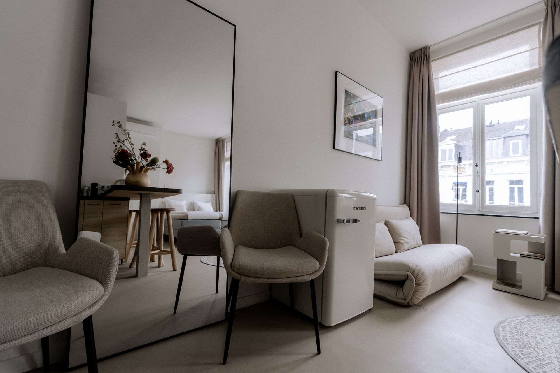 Interieur van een klein appartement met een grote spiegel, stoelen en een bank bij een raam.