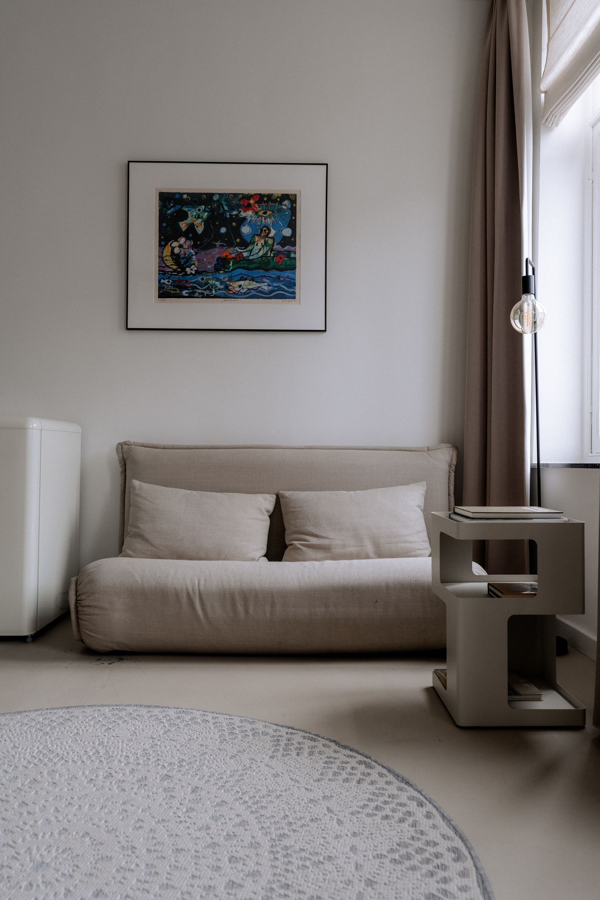 Beige bank met kussens onder een ingelijst schilderij aan een witte muur. Grijs vloerkleed en bijzettafel.