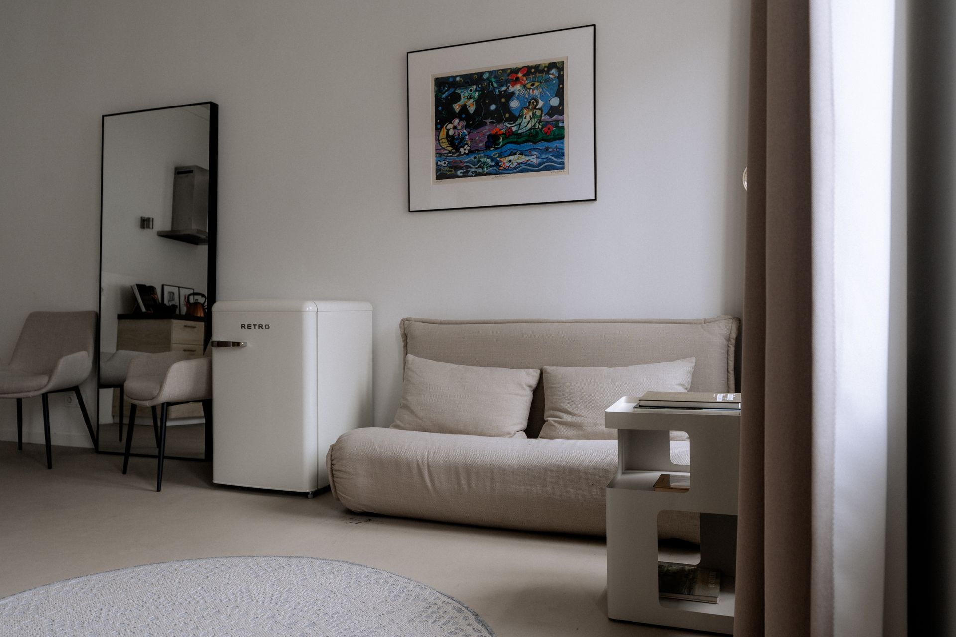 Kamer met neutrale inrichting, bank, schilderij, spiegel, minikoelkast en bijzettafel.