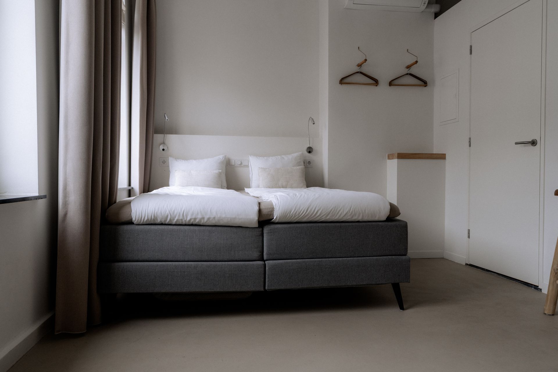 Slaapkamer met een bed, witte muren, grijze gordijnen en twee kledinghangers.
