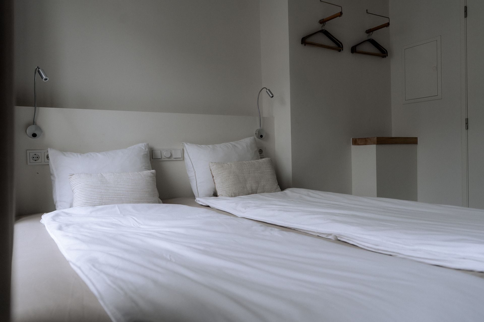 Slaapkamer met twee witte bedden, kussens en lampen tegen een witte muur. Kledinghangers in een kast.