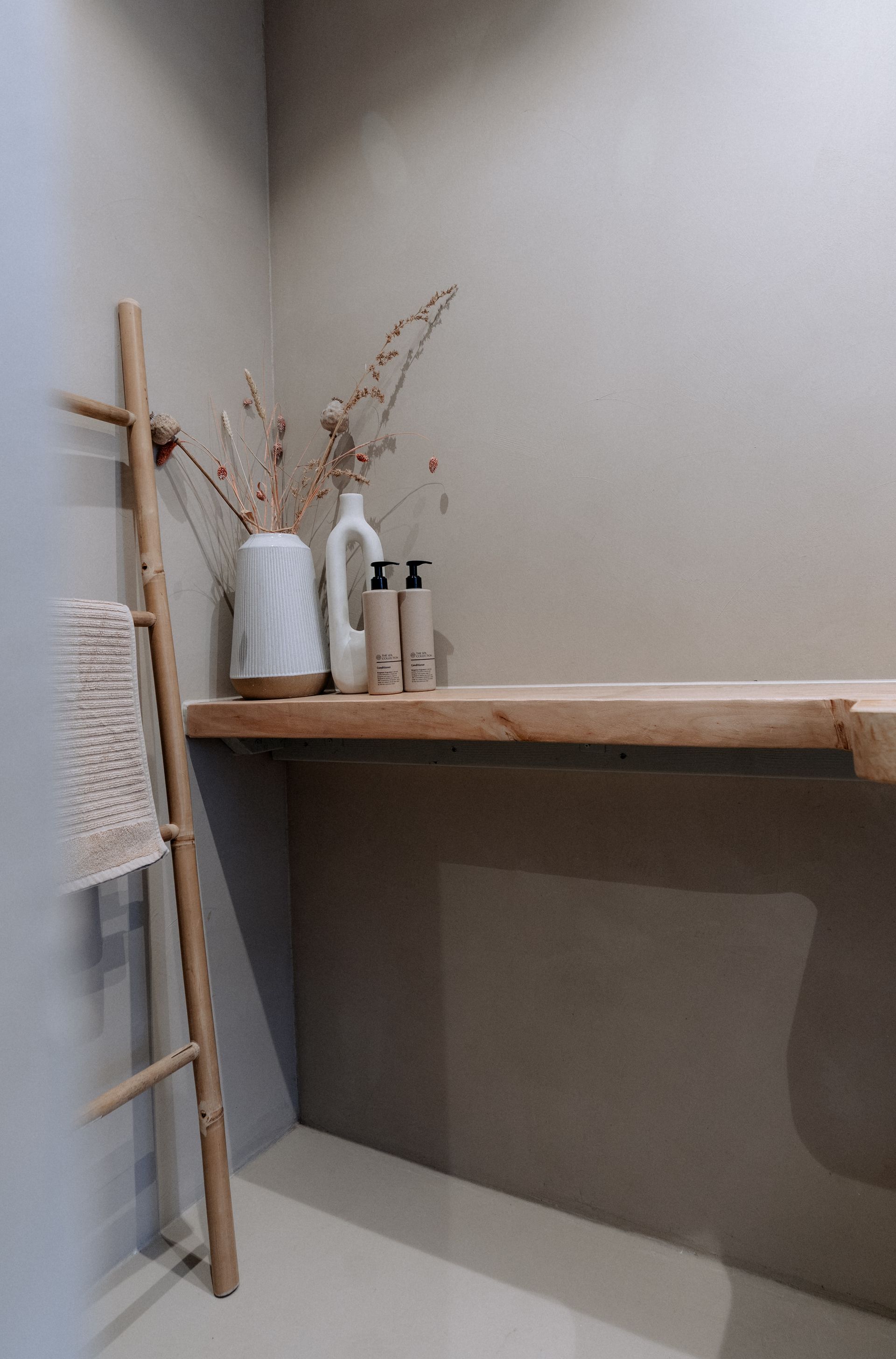 Houten ladder met handdoek naast een plank met vazen ​​en toiletartikelen in een badkamer in neutrale tinten.