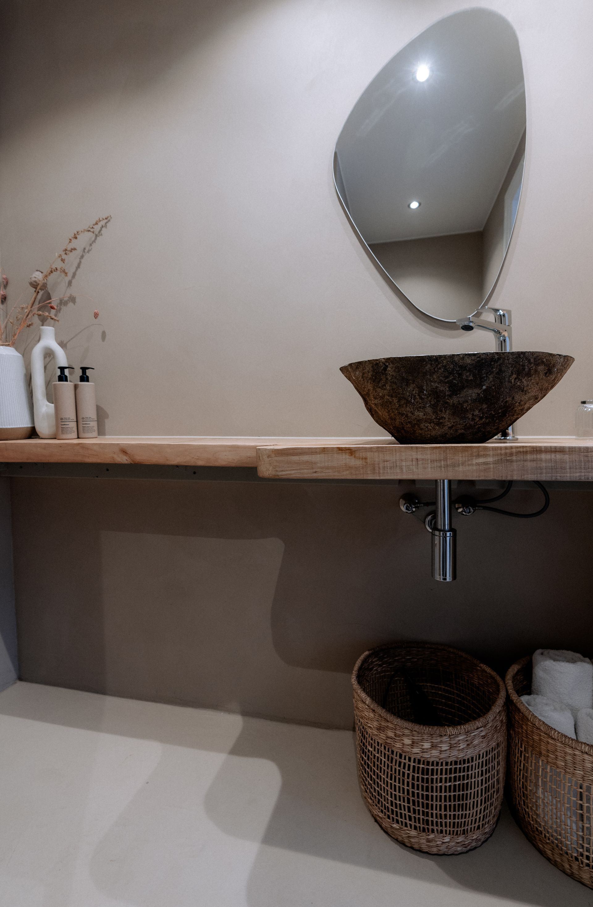 Badkamer met stenen wastafel, spiegel, houten plank en gevlochten manden. Neutrale tinten en natuurlijke texturen.