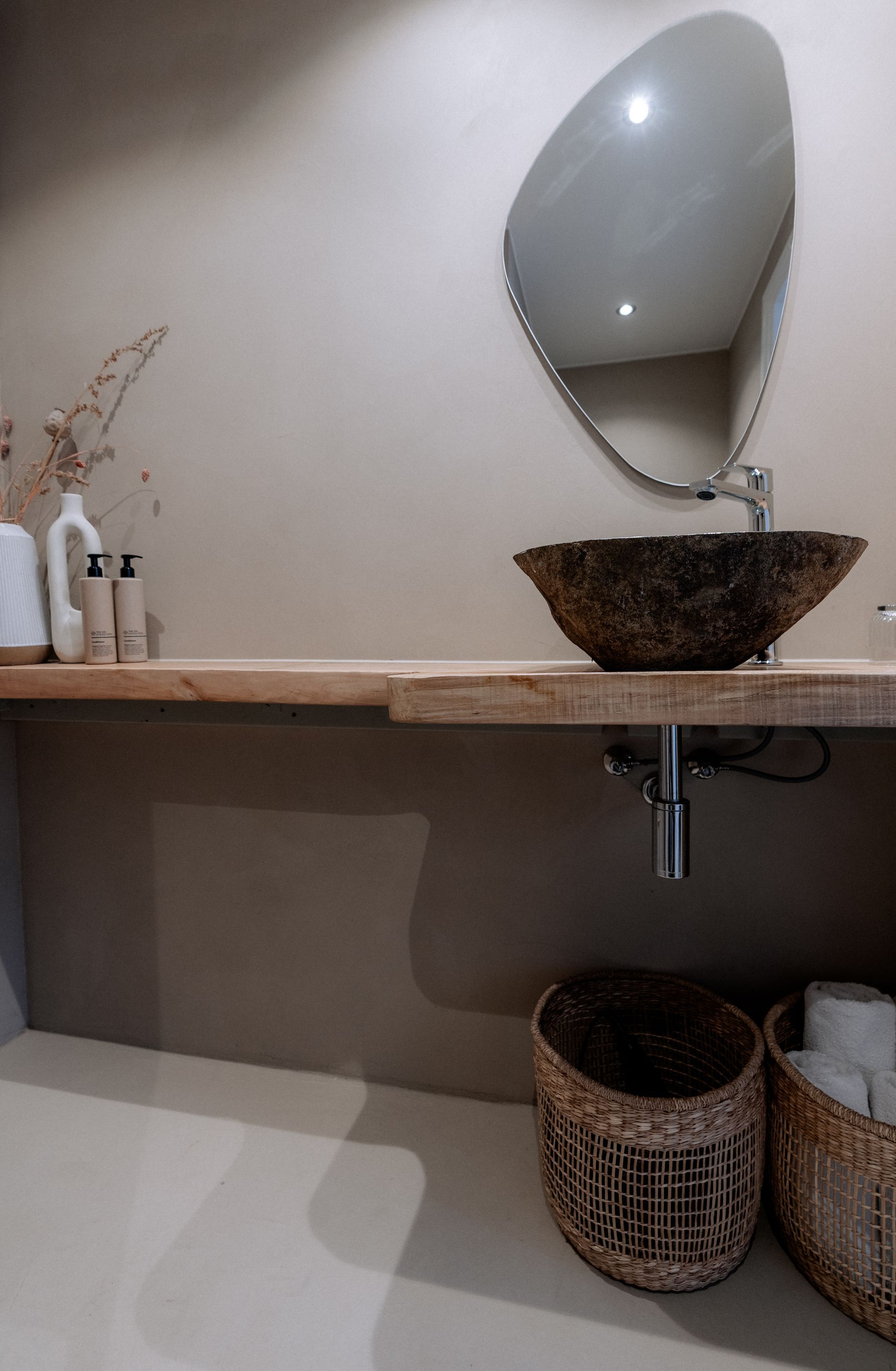 Badkamer met bruine stenen wastafel, ovale spiegel, houten plank en gevlochten manden.