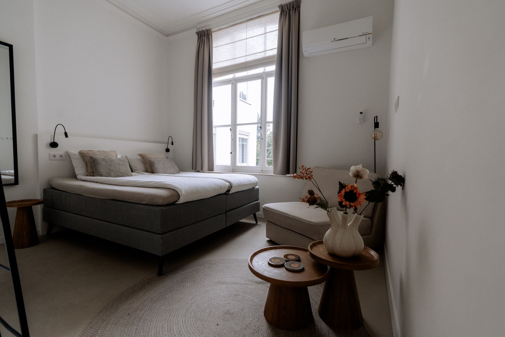 Slaapkamer met een bed, fauteuil, nachtkastjes, raam en spiegel. Witte muren en zachte grijs- en beigetinten.