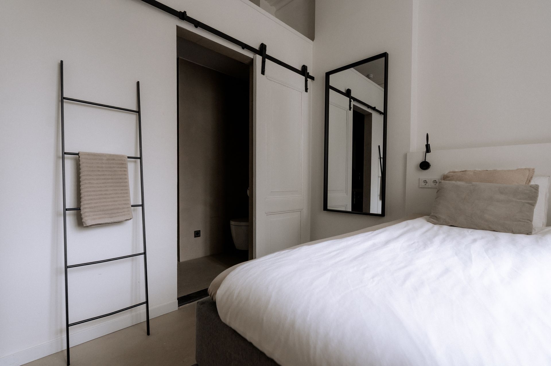 Slaapkamer met wit beddengoed, schuifdeur naar de badkamer en een decoratieve ladder met een handdoek.