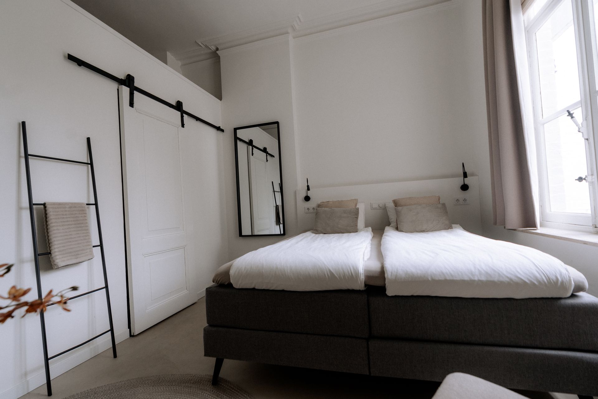 Witte slaapkamer met een bed, schuifdeur, spiegel en een ladder tegen de muur.