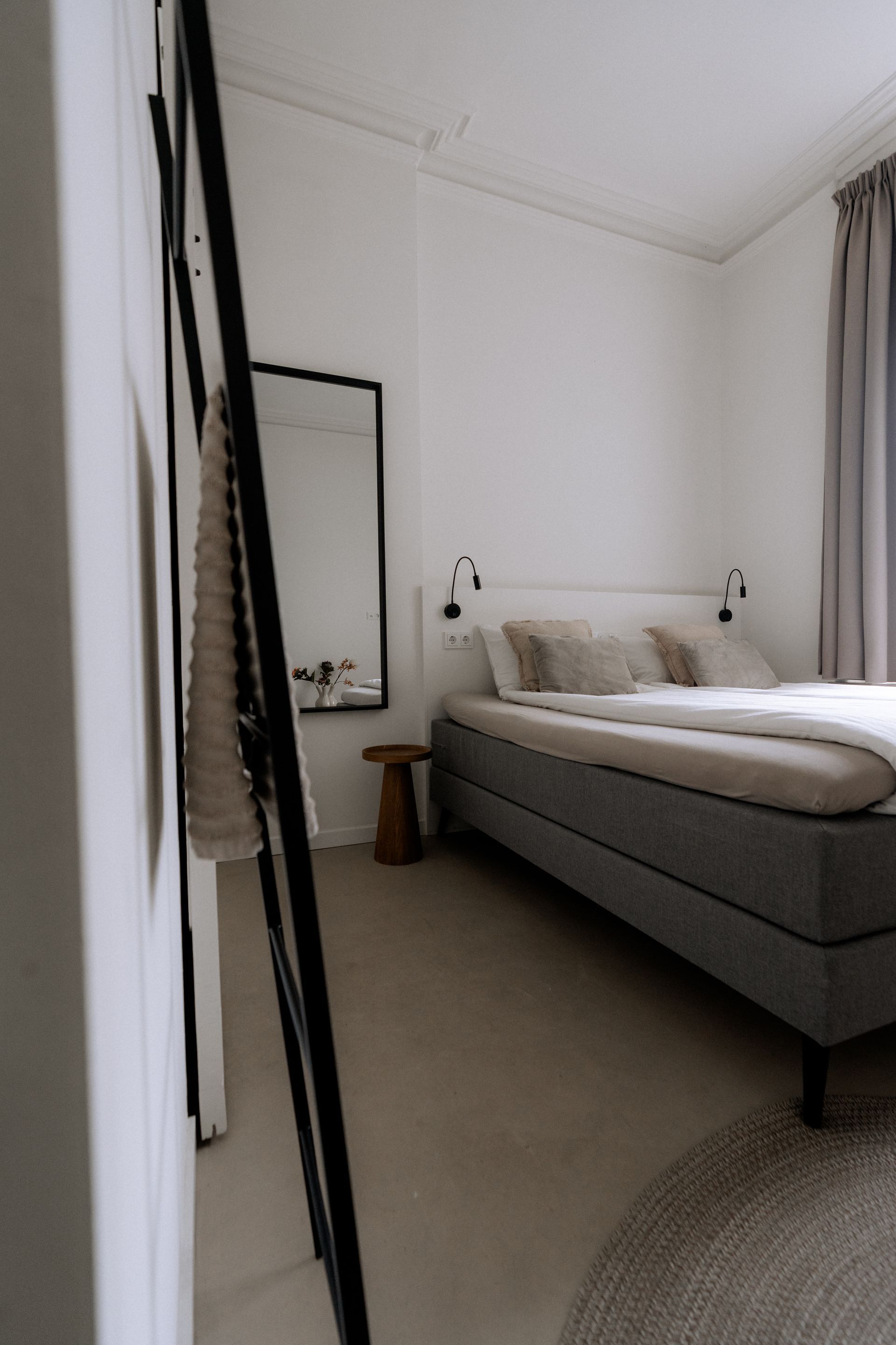 Slaapkamerinterieur met een bed, spiegel en een klein krukje. Witte muren, grijs beddengoed en neutrale tinten.