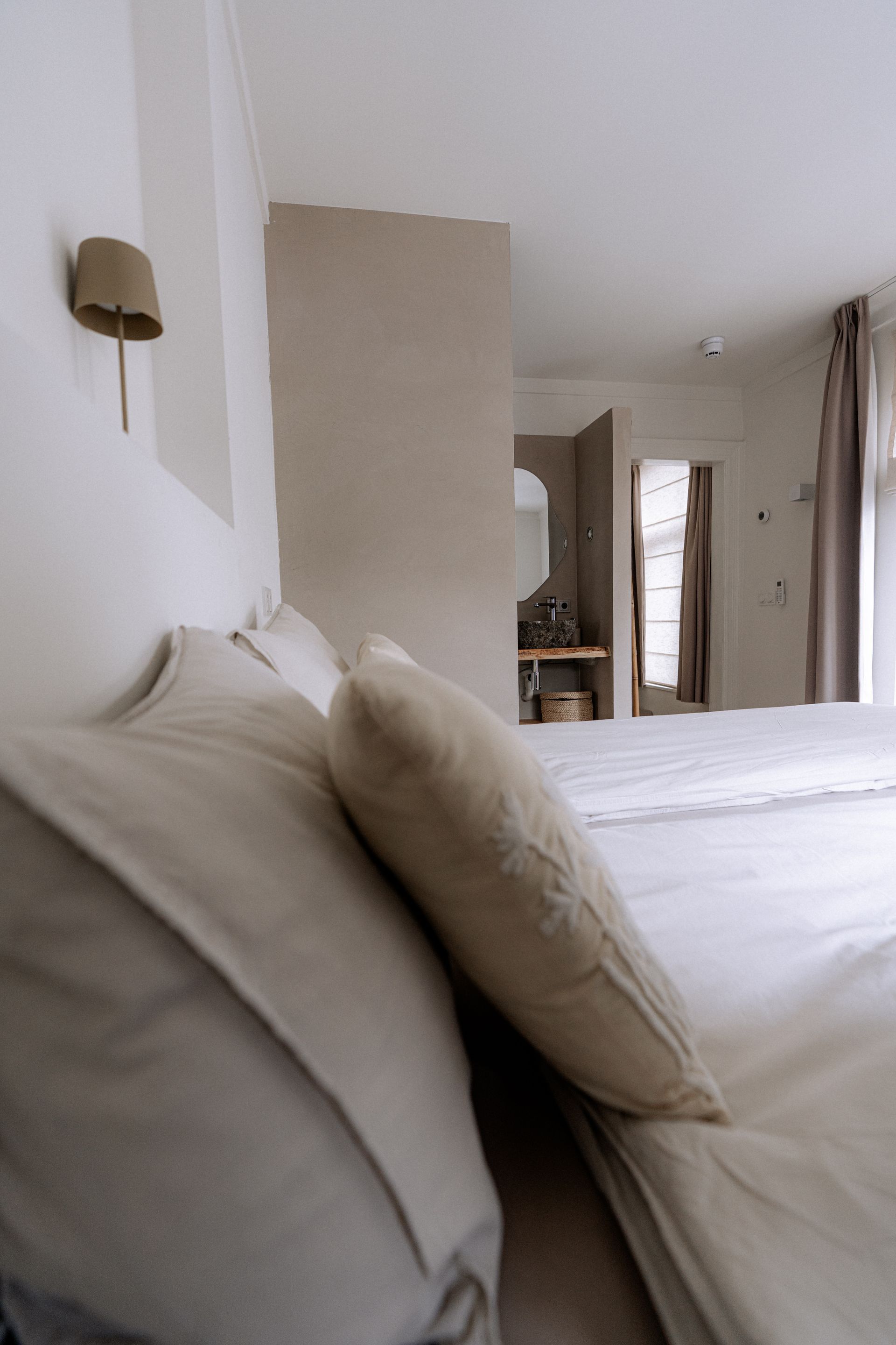Een bed met wit beddengoed en lichtgekleurde kussens. Uitzicht op de badkamer.