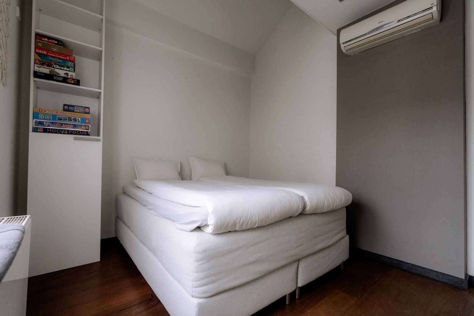 Kleine, moderne slaapkamer met wit bed, ingebouwde planken, airconditioning en donkere houten vloer.