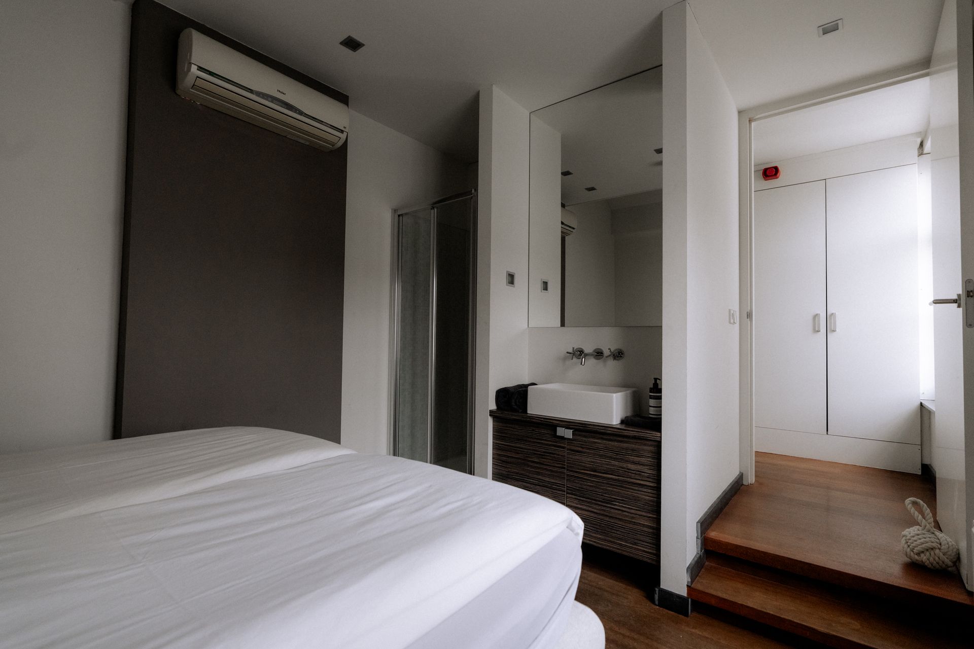 Slaapkamer met een bed, wastafel en een deur naar de trap. Witte muren, houten vloer.