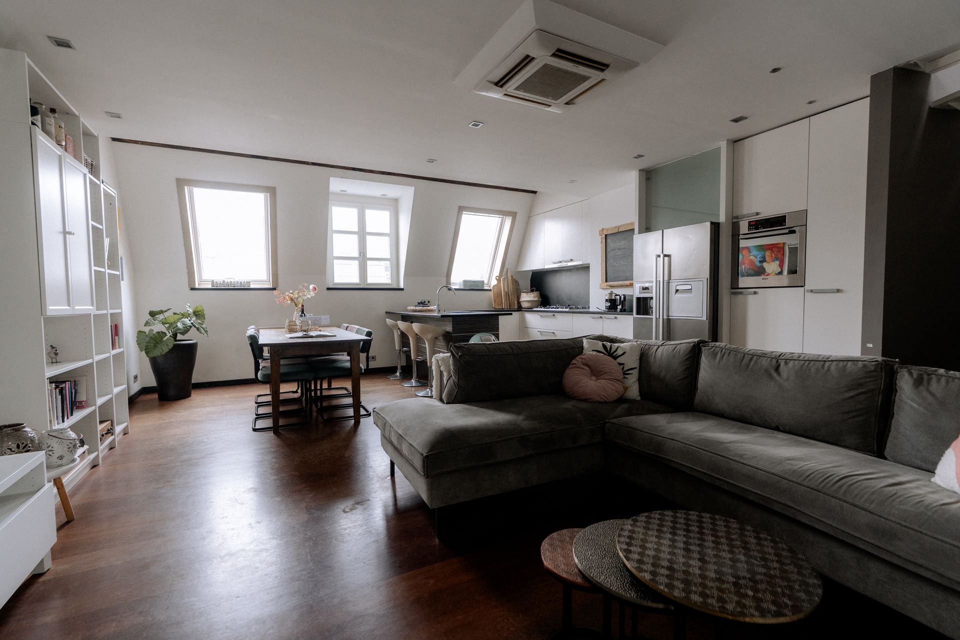 Woonkamer met donkere houten vloer, grote grijze bank, eettafel en keukenhoek.