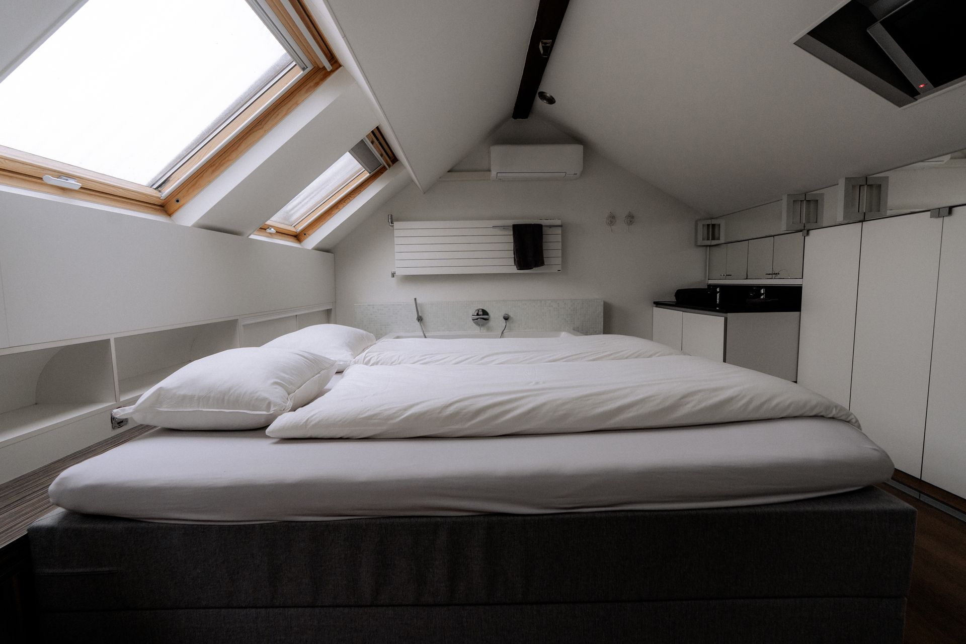 Slaapkamer met bed onder een schuin plafond, dakraam erboven, witte muren en ingebouwde opbergruimte.