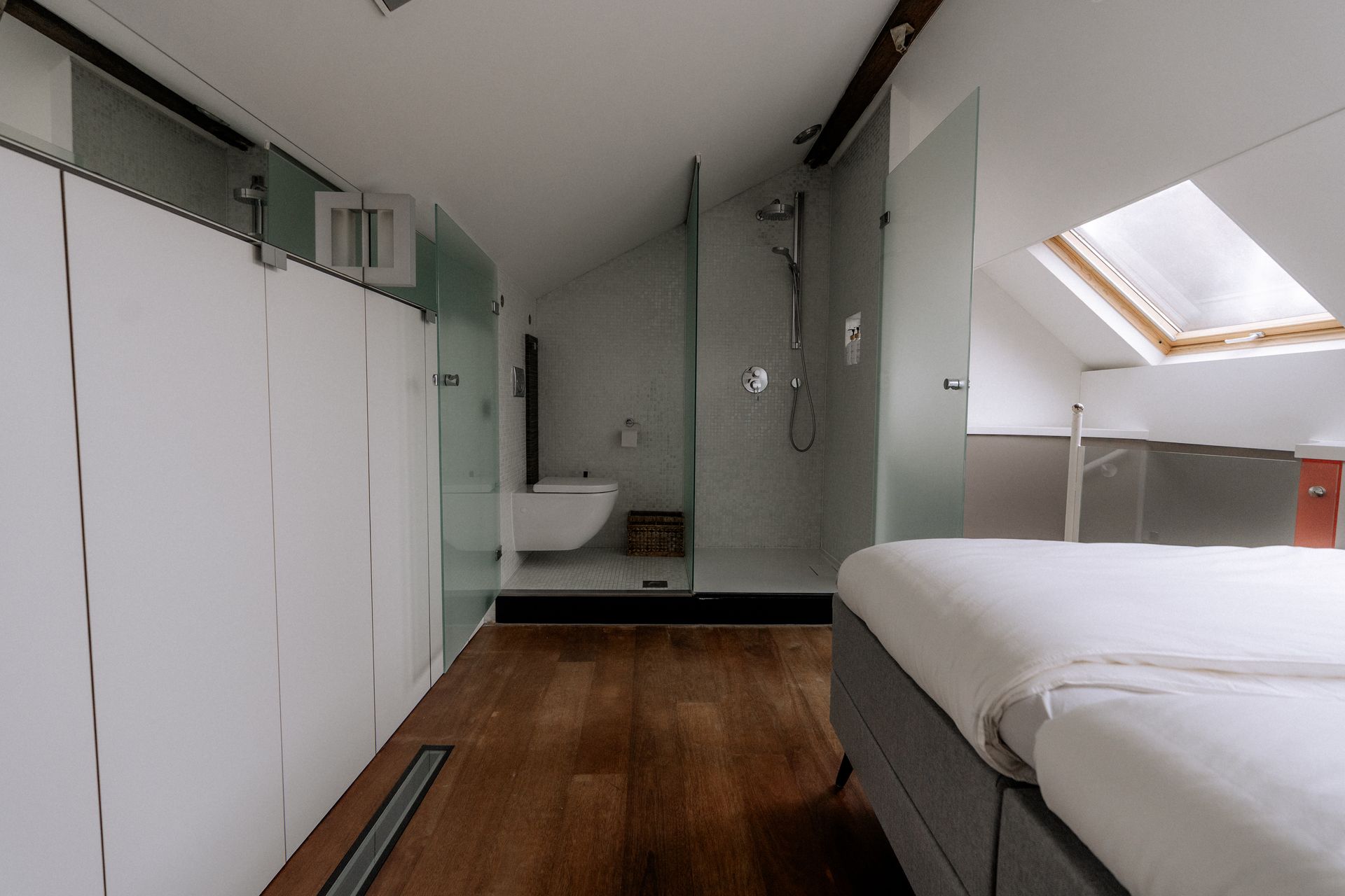 Moderne badkamer met een bed op de voorgrond, douche, toilet en dakraam. Houten vloer, witte muren.