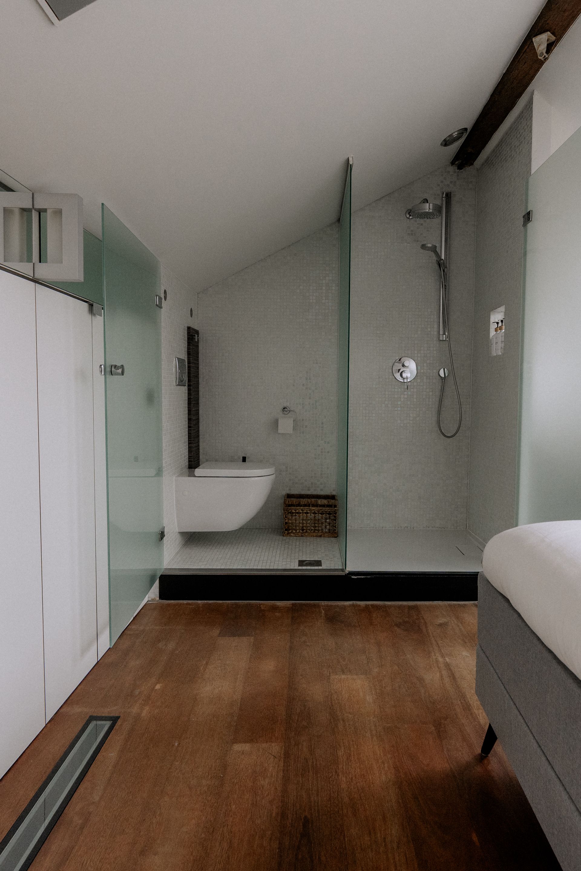 Badkamer met houten vloer, glazen douchecabine, wit toilet en zichtbare balken.