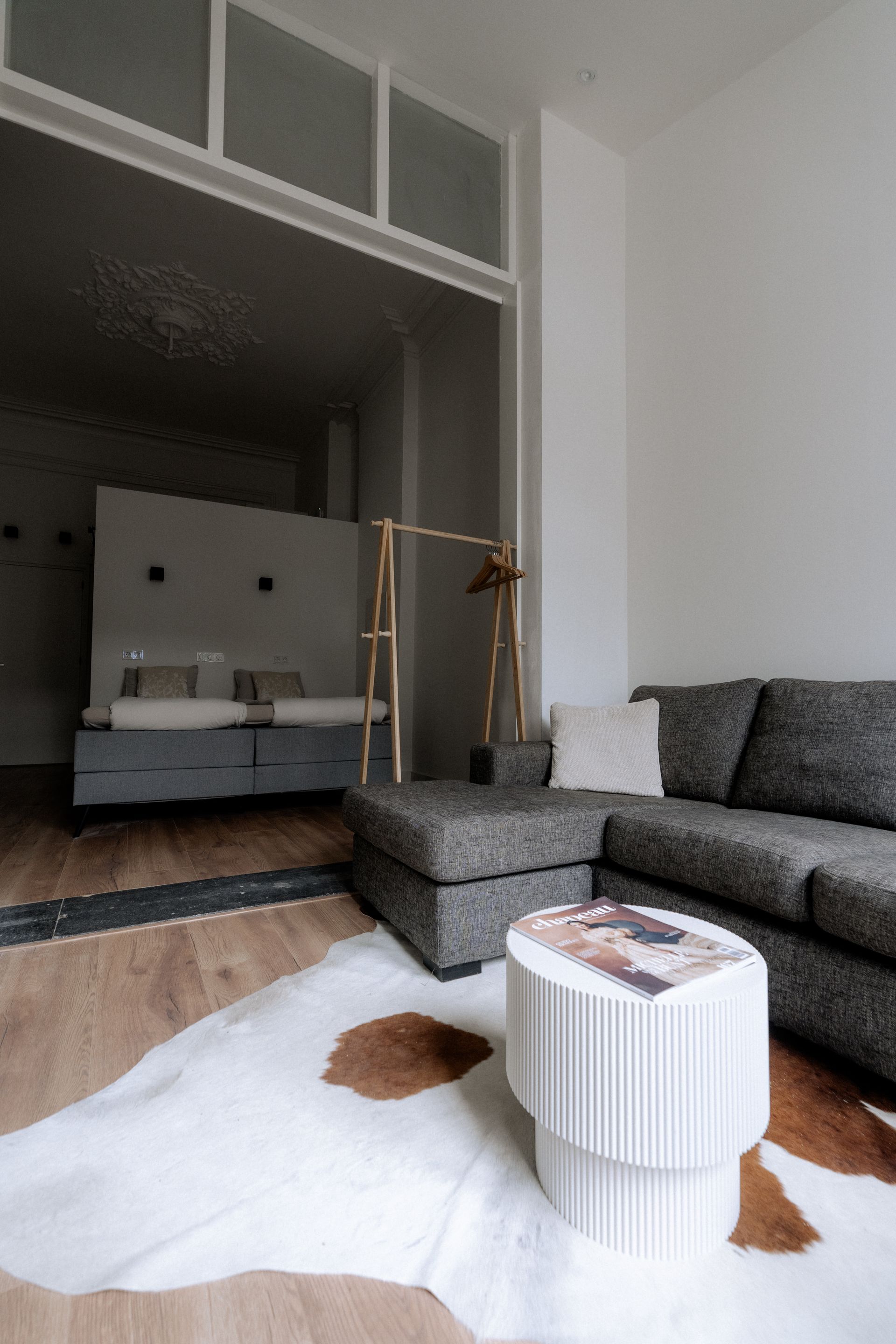 Woonkamer met grijze bank, poef en witte salontafel op een koeienhuid vloerkleed, met uitzicht op de slaapkamer.