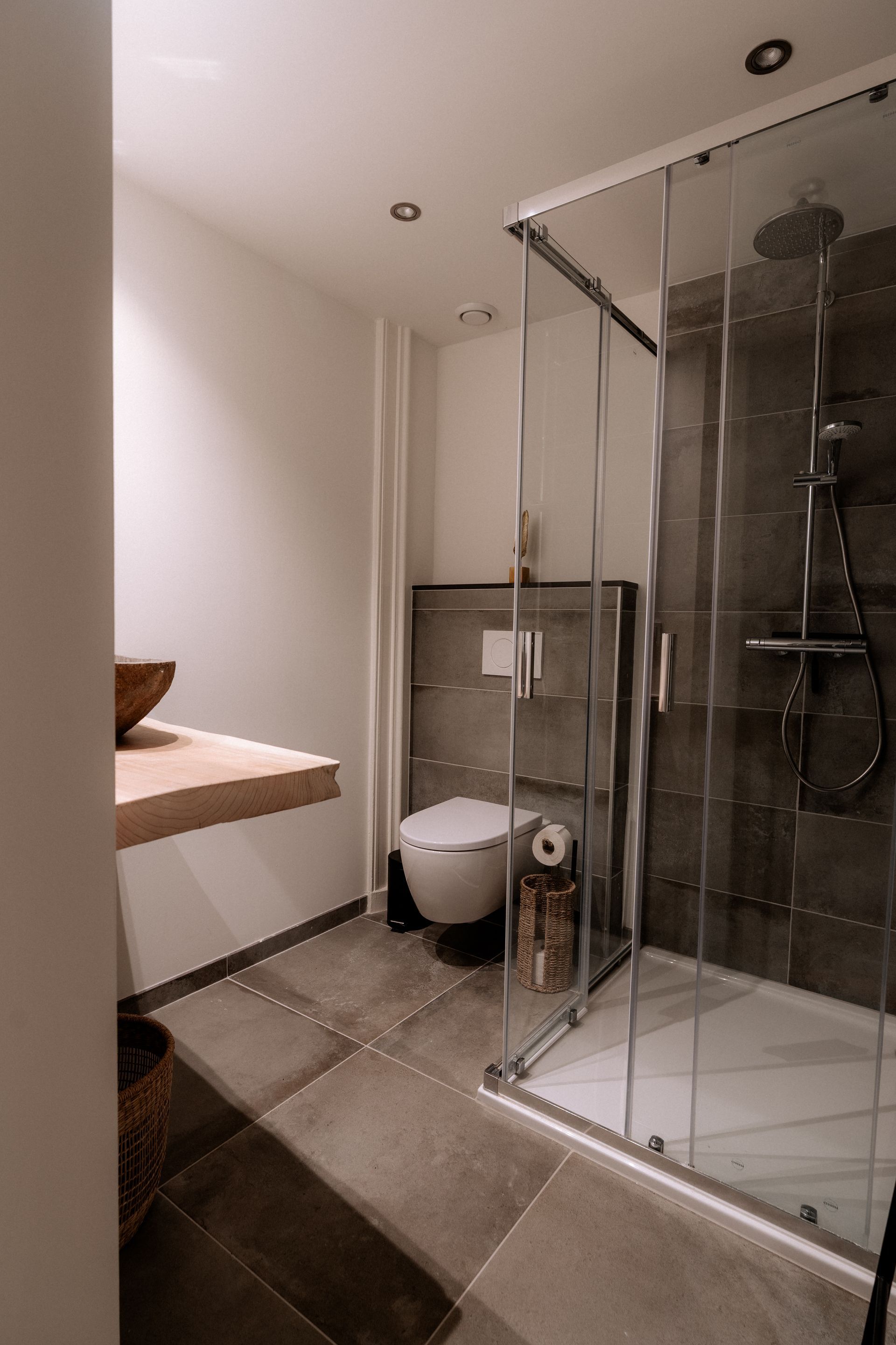 Moderne badkamer met inloopdouche, toilet en houten aanrechtblad met wastafel. Grijze tegelvloer en -wanden.