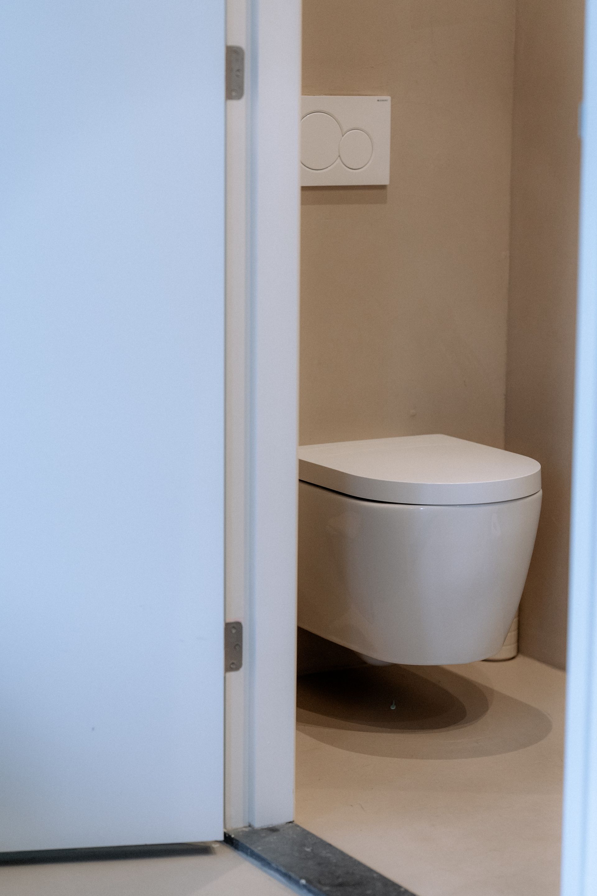 Wit, aan de muur gemonteerd toilet in een kleine badkamer, zichtbaar door een gedeeltelijk openstaande deur. Beige muren.