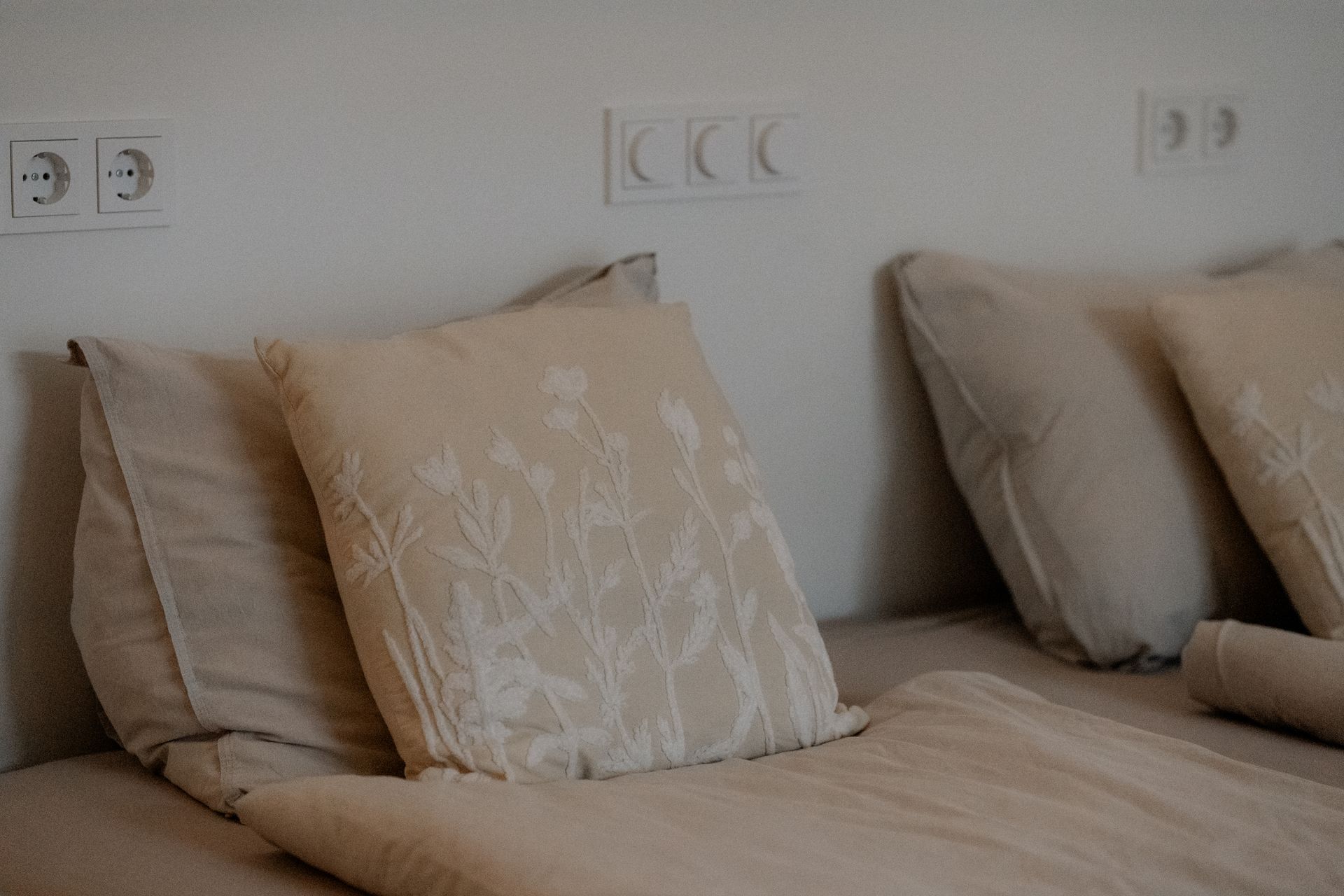 Beige kussens en beddengoed op een bed tegen een witte muur met stopcontacten.
