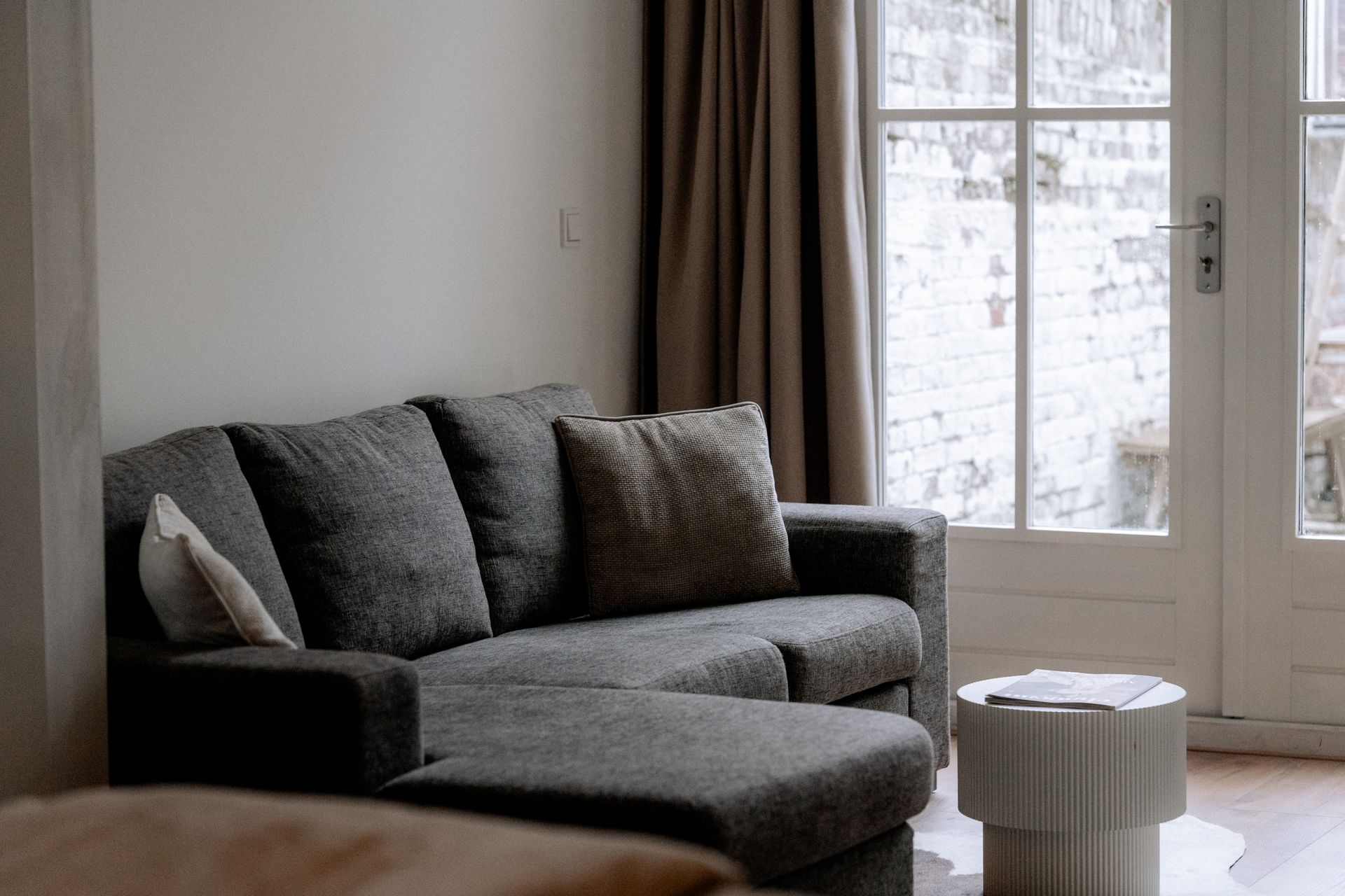 Grijze bank met chaise longue in een kamer, naast een raam met een klein tafeltje.