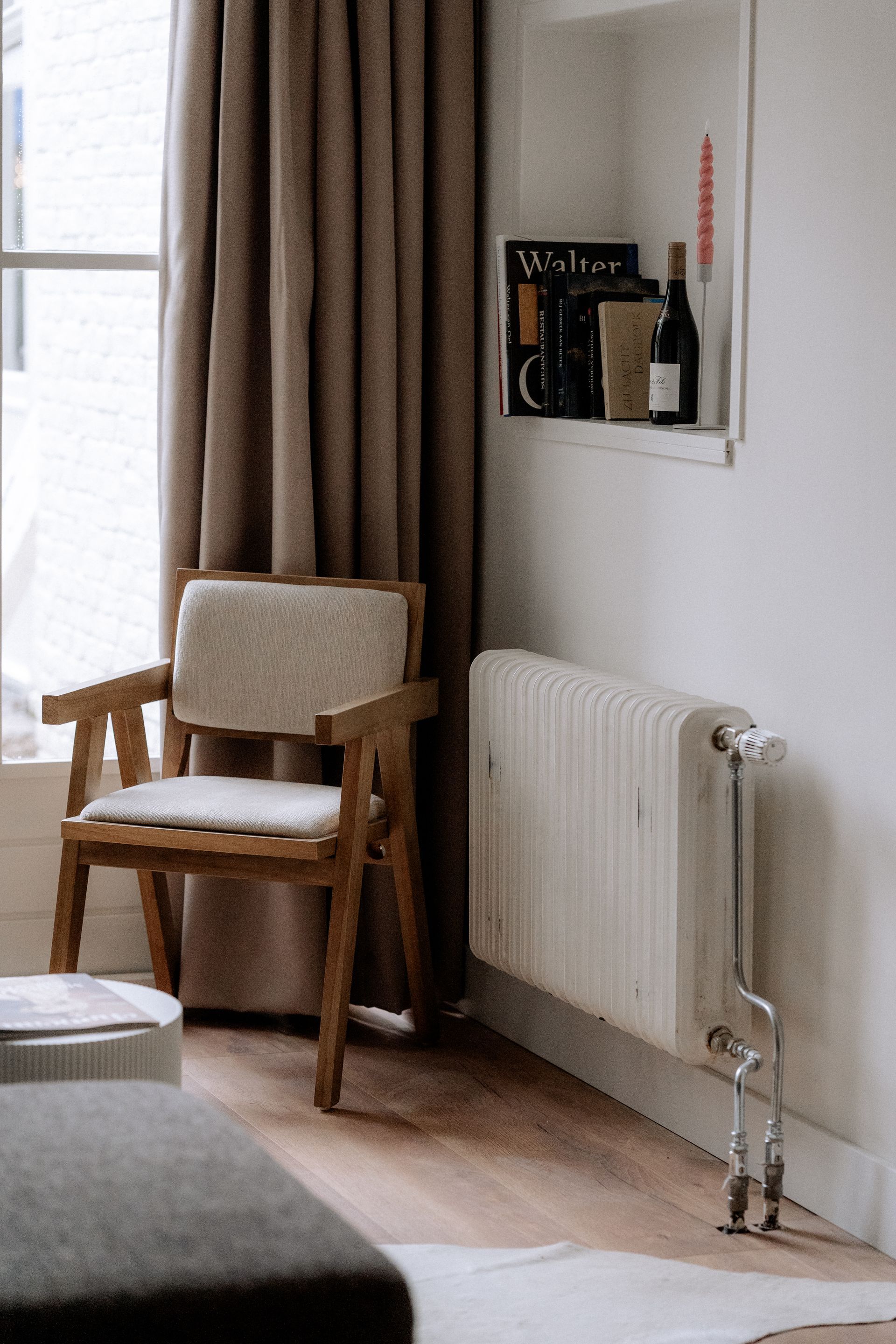 Houten stoel bij een raam met bruine gordijnen. Witte muur, radiator, boeken en een fles op een plank.