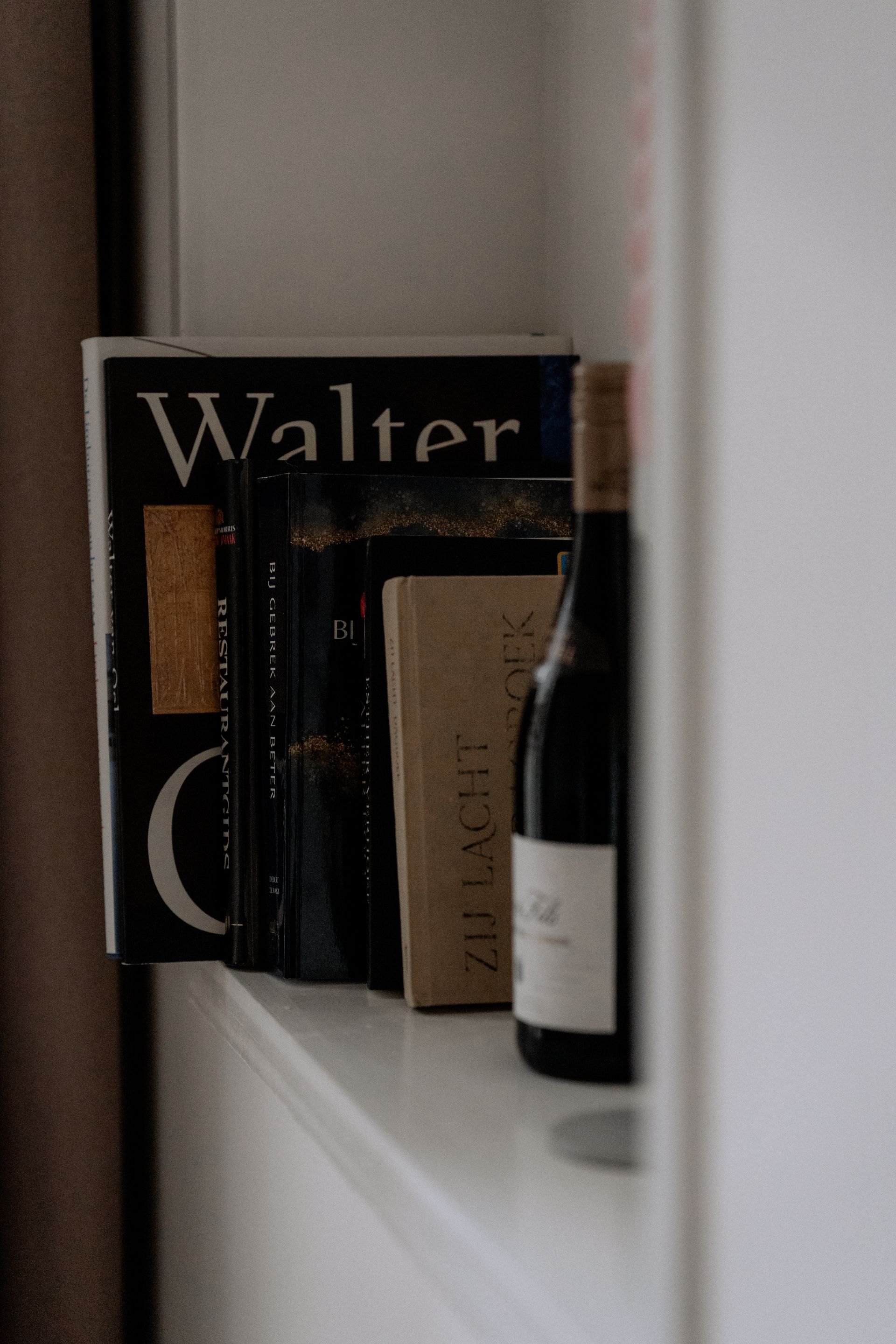 Boeken en een wijnfles op een witte plank. De boeken zijn donkerbruin en lichtbruin. De wijnfles is donker.