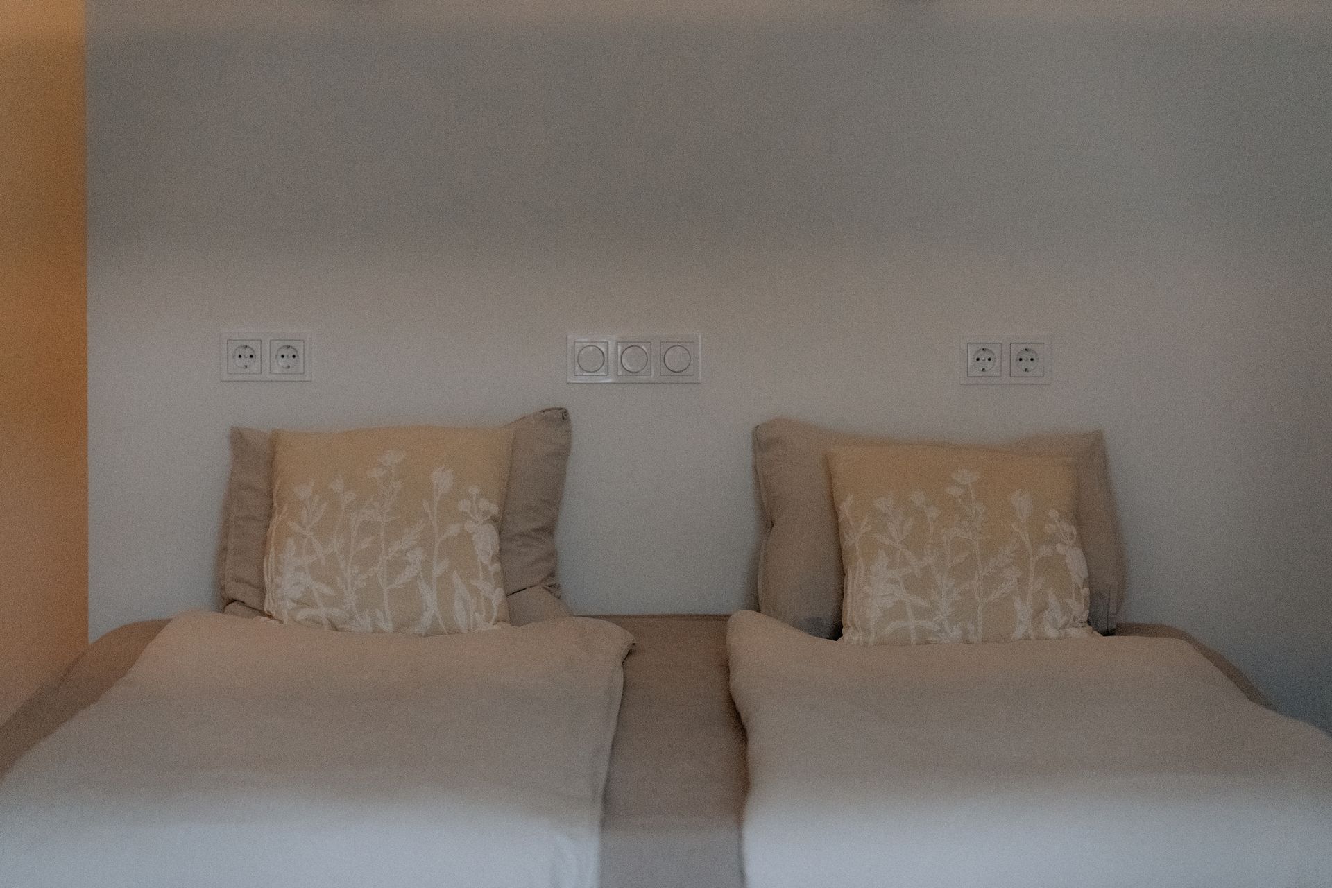 Twee bedden met wit beddengoed en sierkussens tegen een witte muur met stopcontacten.